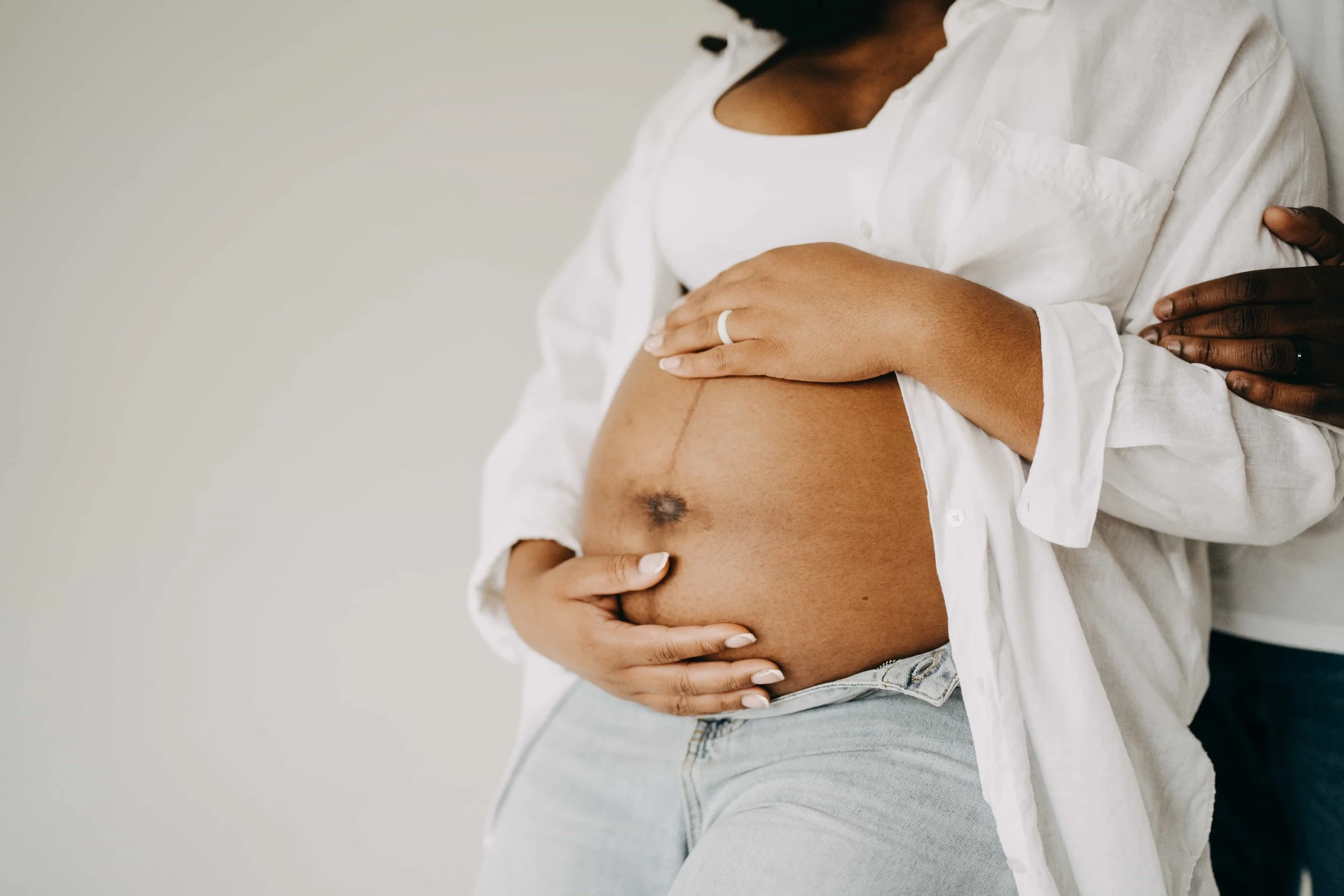 A pregnant woman in a white shirt and jeans is gently holding her baby bump. A person behind her has their hand on her shoulder, showing support.