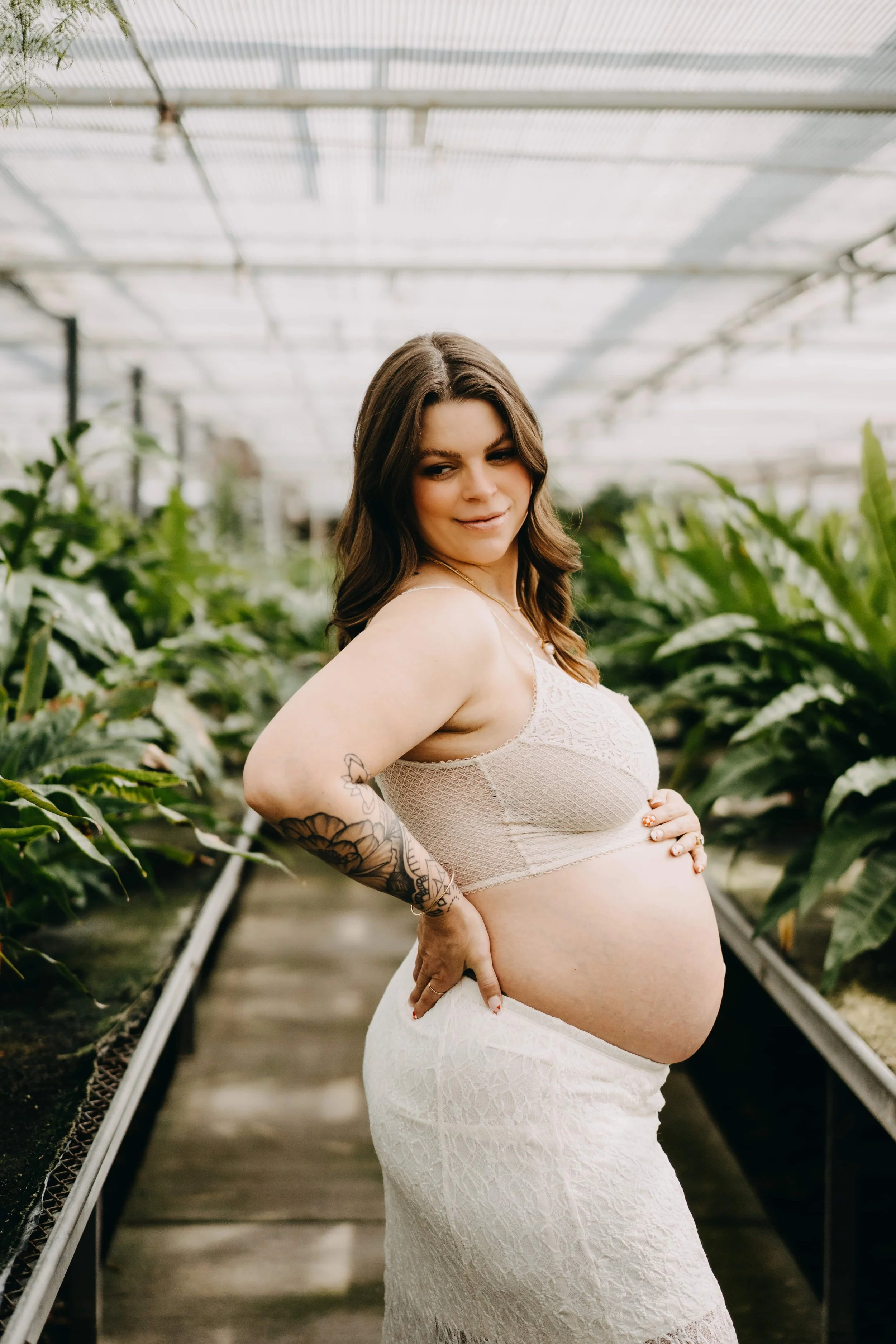 A pregnant woman with dark hair and tattoos on her left arm stands in a greenhouse, wearing a lace top and a white skirt, with plants on either side.