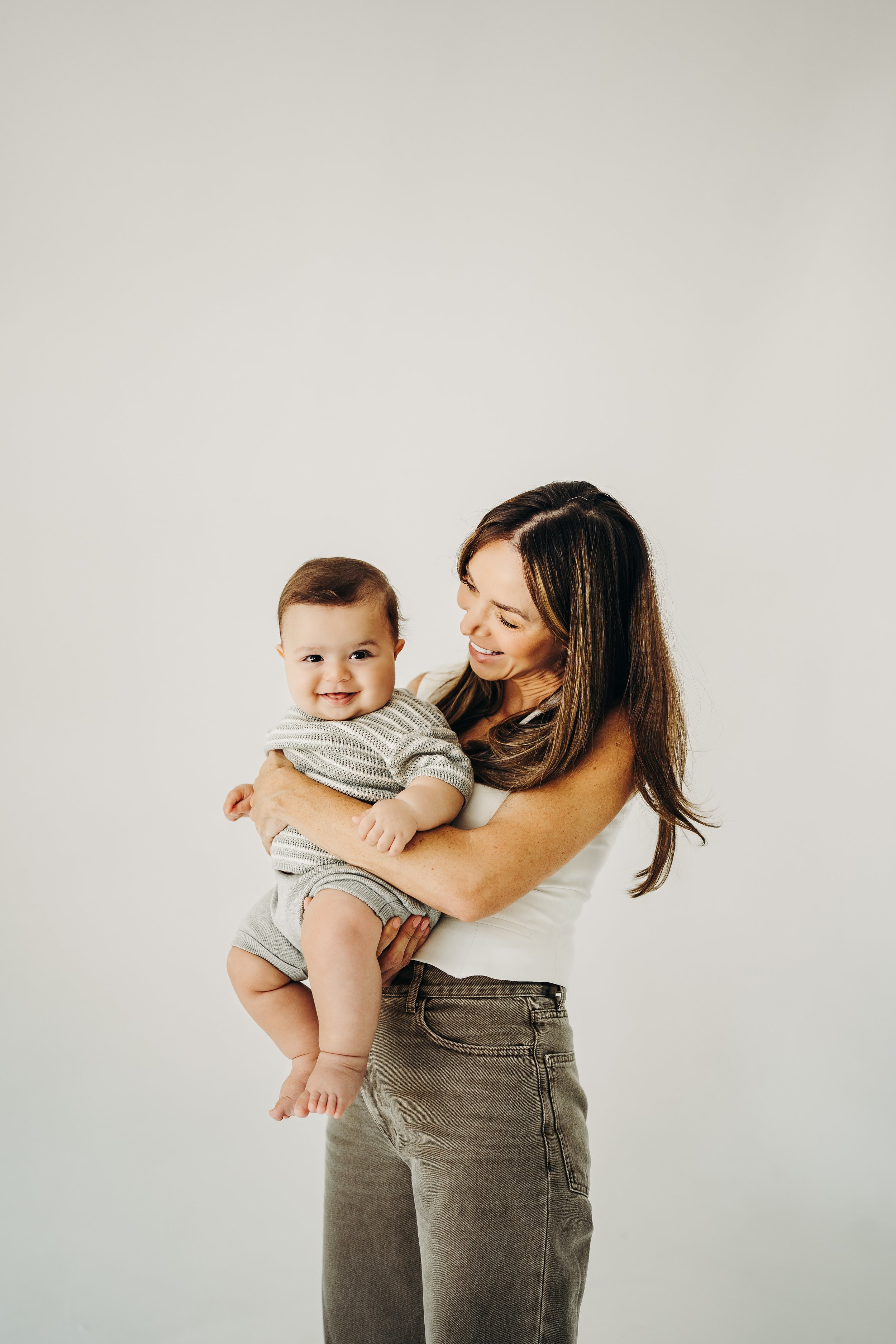 Woman smiling and holding a baby in grey outfit against a plain background.
