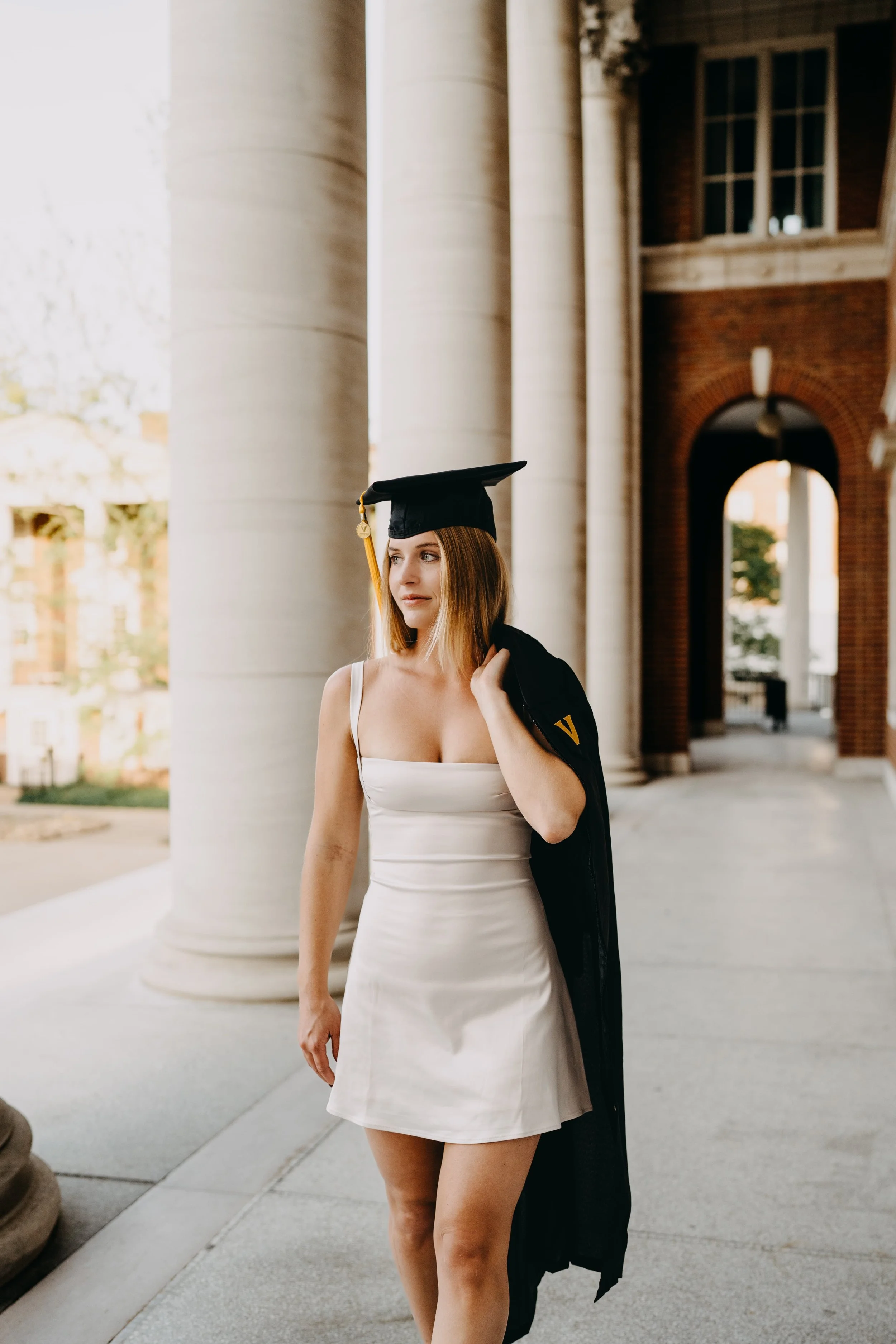 A young woman in a white dress and graduation cap standing outside near large white columns of a building, holding a black jacket over her shoulder. She has shoulder-length hair and is looking slightly to the side.