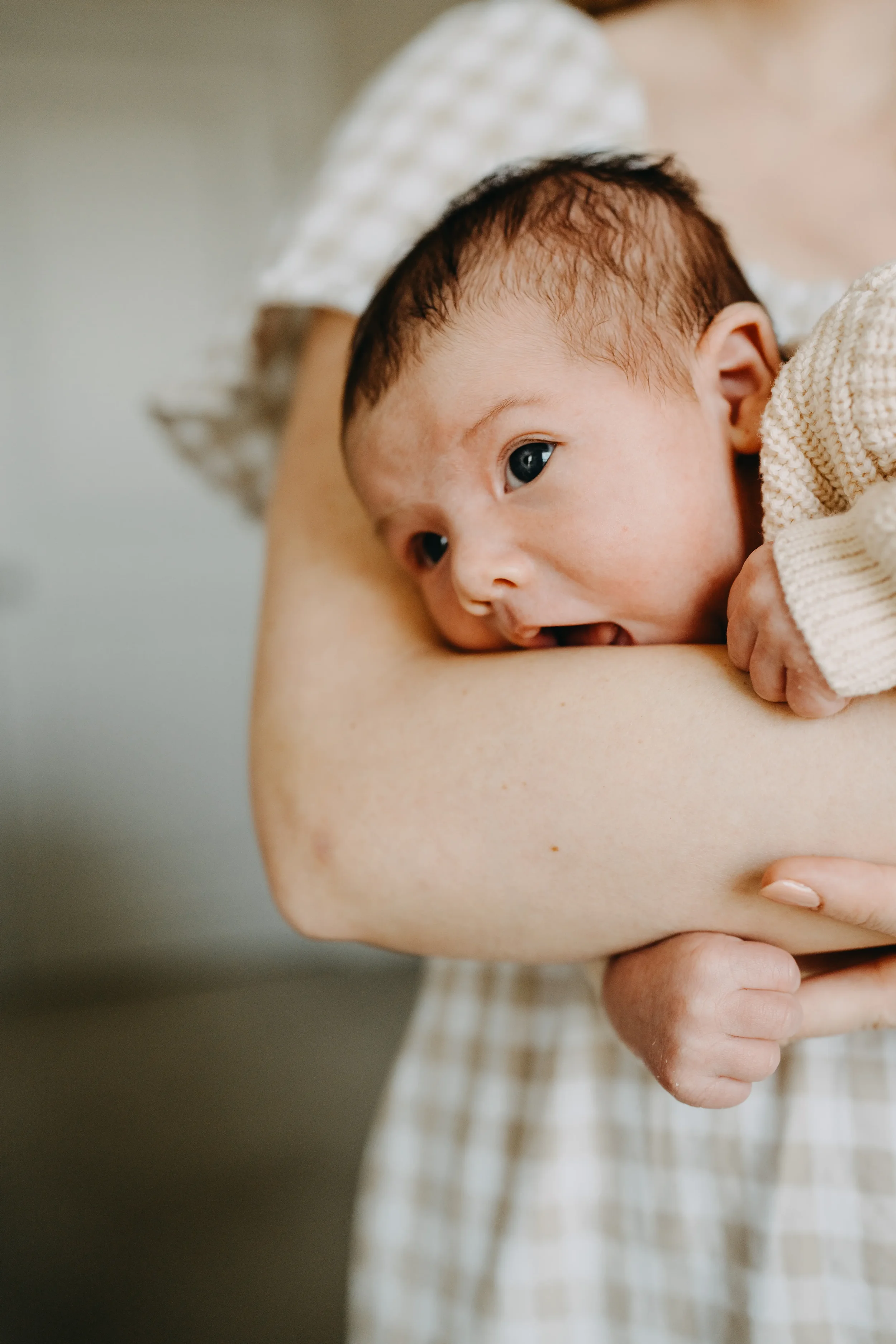 A close-up of a baby lying on someone's arm, resting on a person's shoulder. The baby has dark hair, big eyes, and is wearing a beige knitted garment.