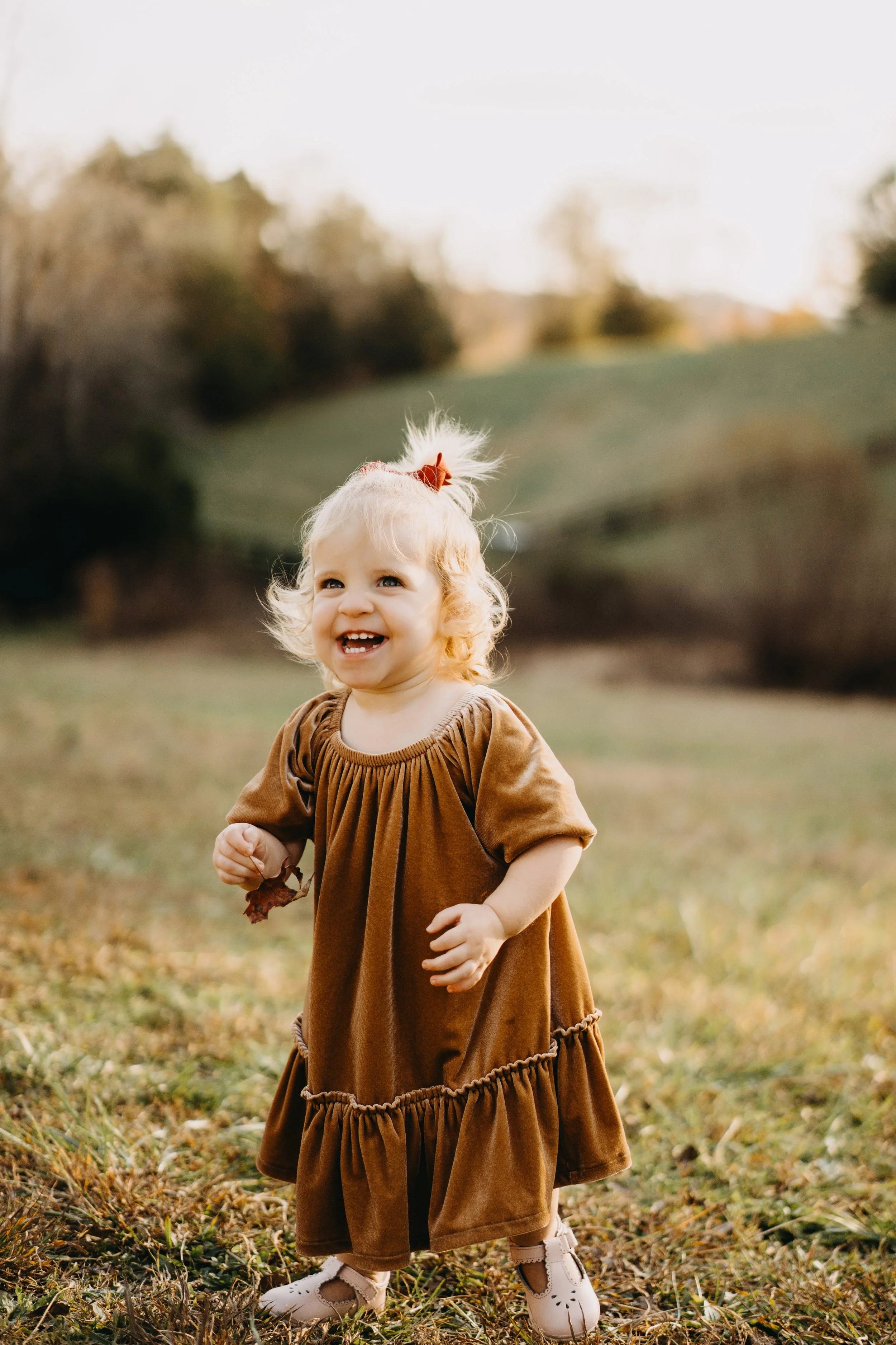A young girl with curly blonde hair, wearing a brown dress with ruffles, is standing outdoors on grass with trees in the background, smiling and holding autumn leaves.
