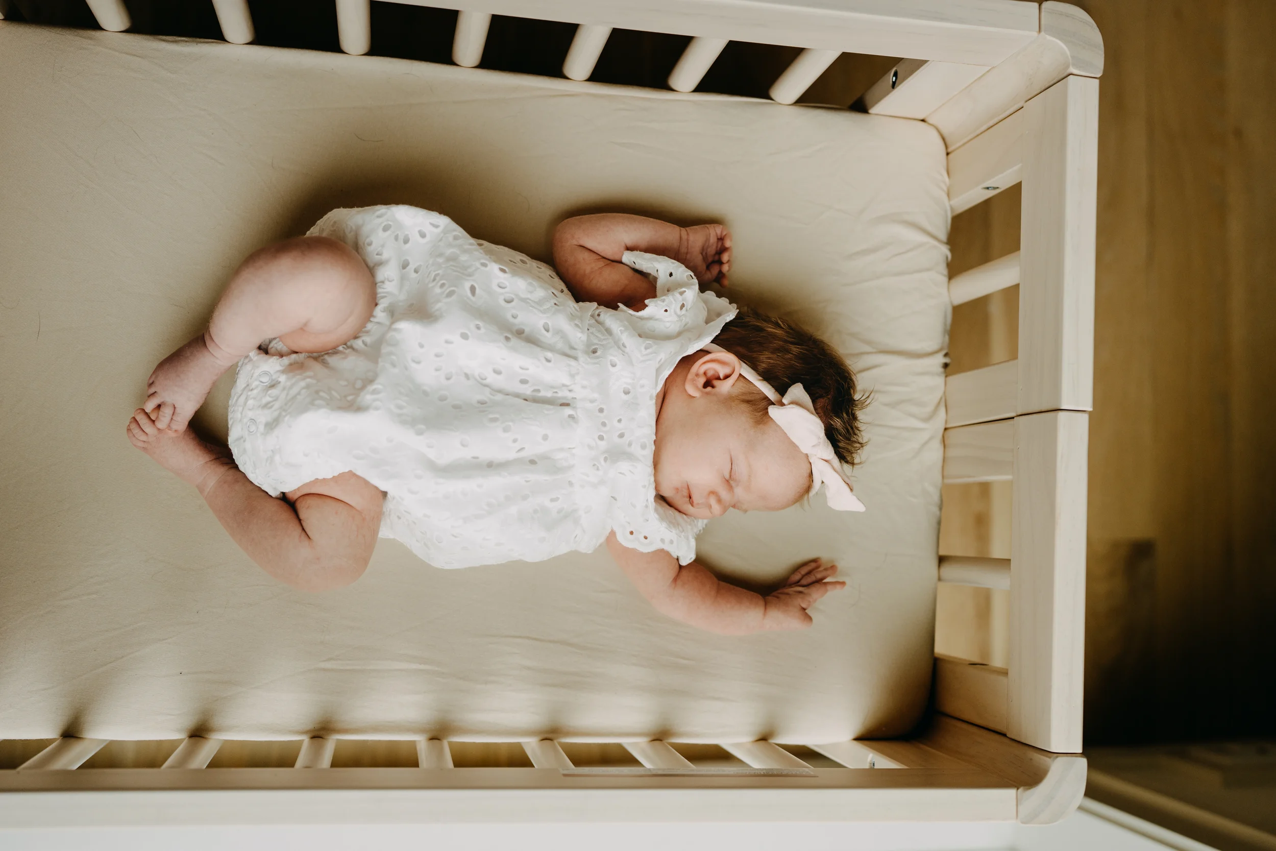 A sleeping infant with brown hair, dressed in a white eyelet dress with ruffled sleeves, lying on their side in a wooden crib with vertical slats and a beige mattress.