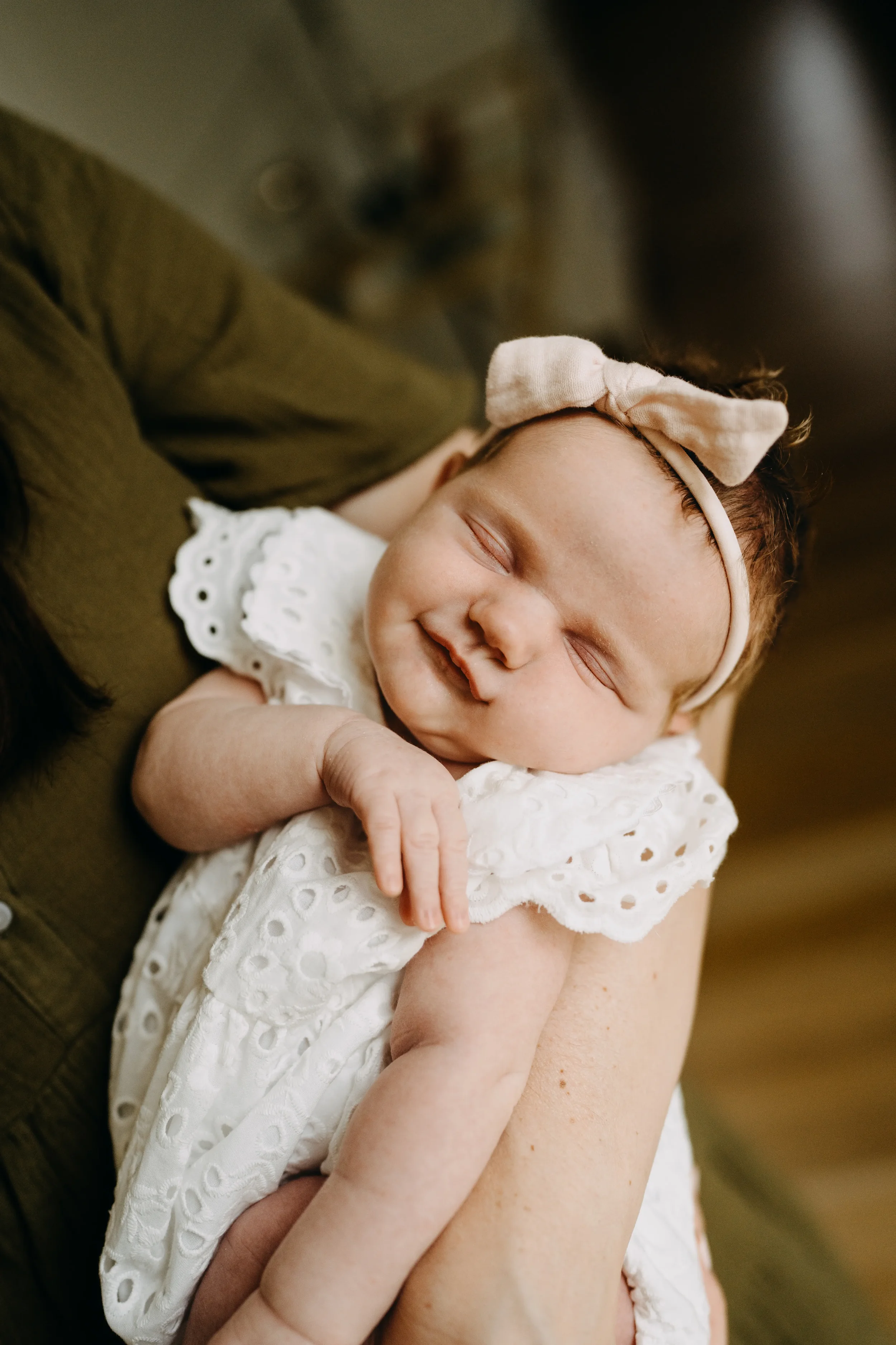 A baby girl sleeping peacefully, wearing a cream-colored headband with a bow and a white eyelet dress, cradled in someone's arm.