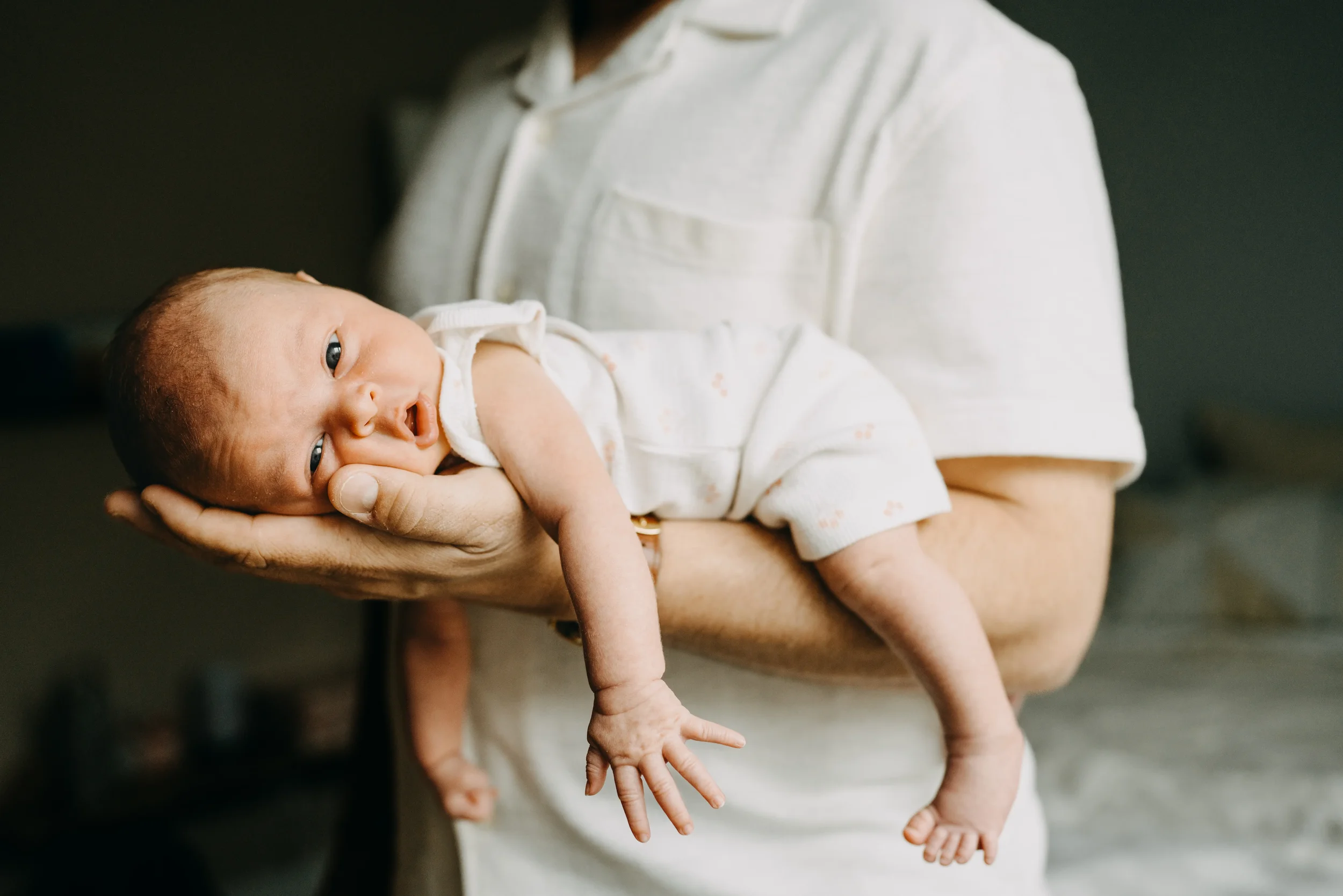 A person holding a newborn baby close to their chest. The baby has light skin, blue eyes, and is dressed in a white outfit with small dots. The adult's face is not visible, and they are wearing a light-colored shirt.