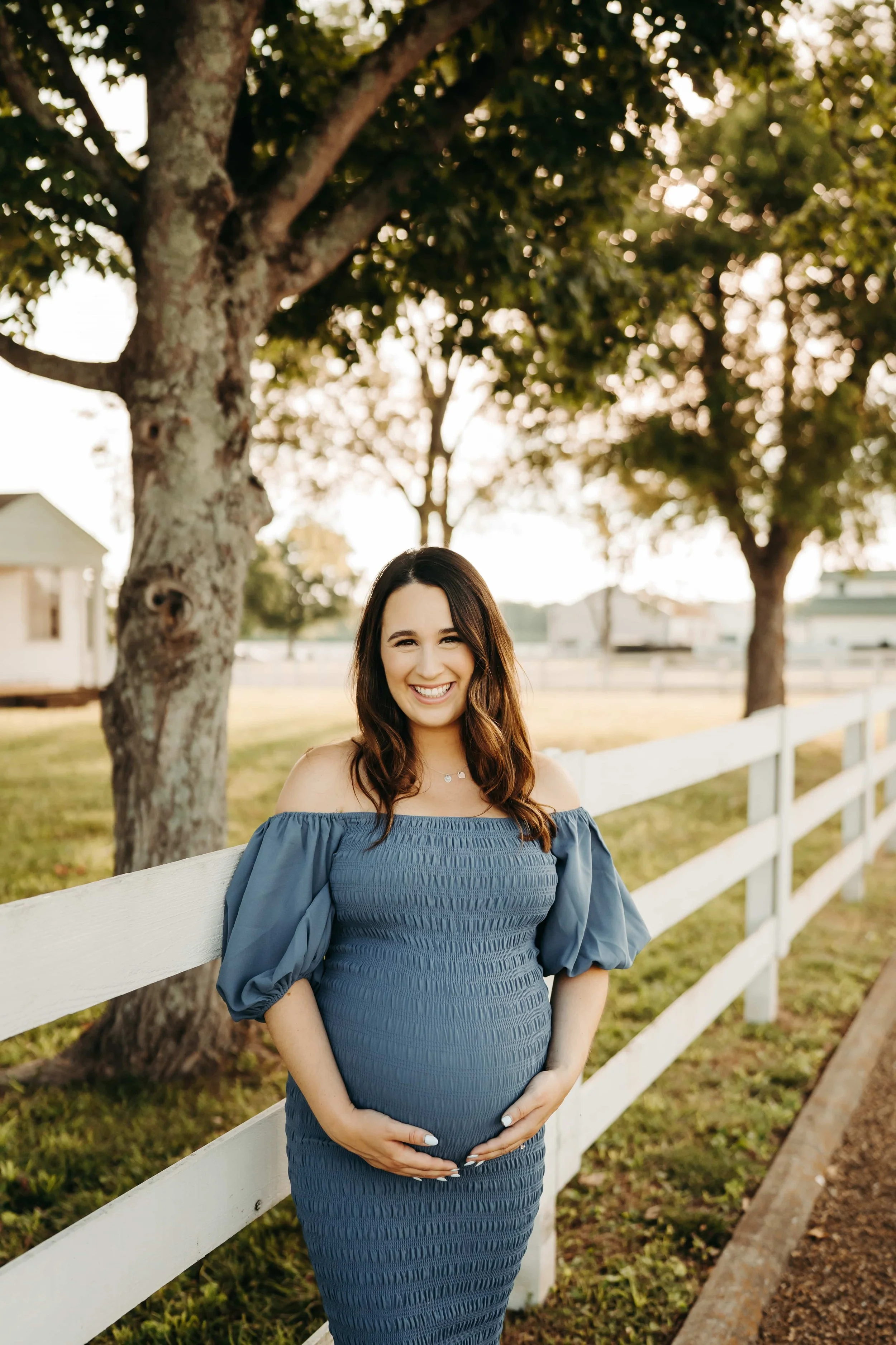 Smiling pregnant woman standing outdoors by a white fence, holding her belly, with trees and houses in the background during sunset.