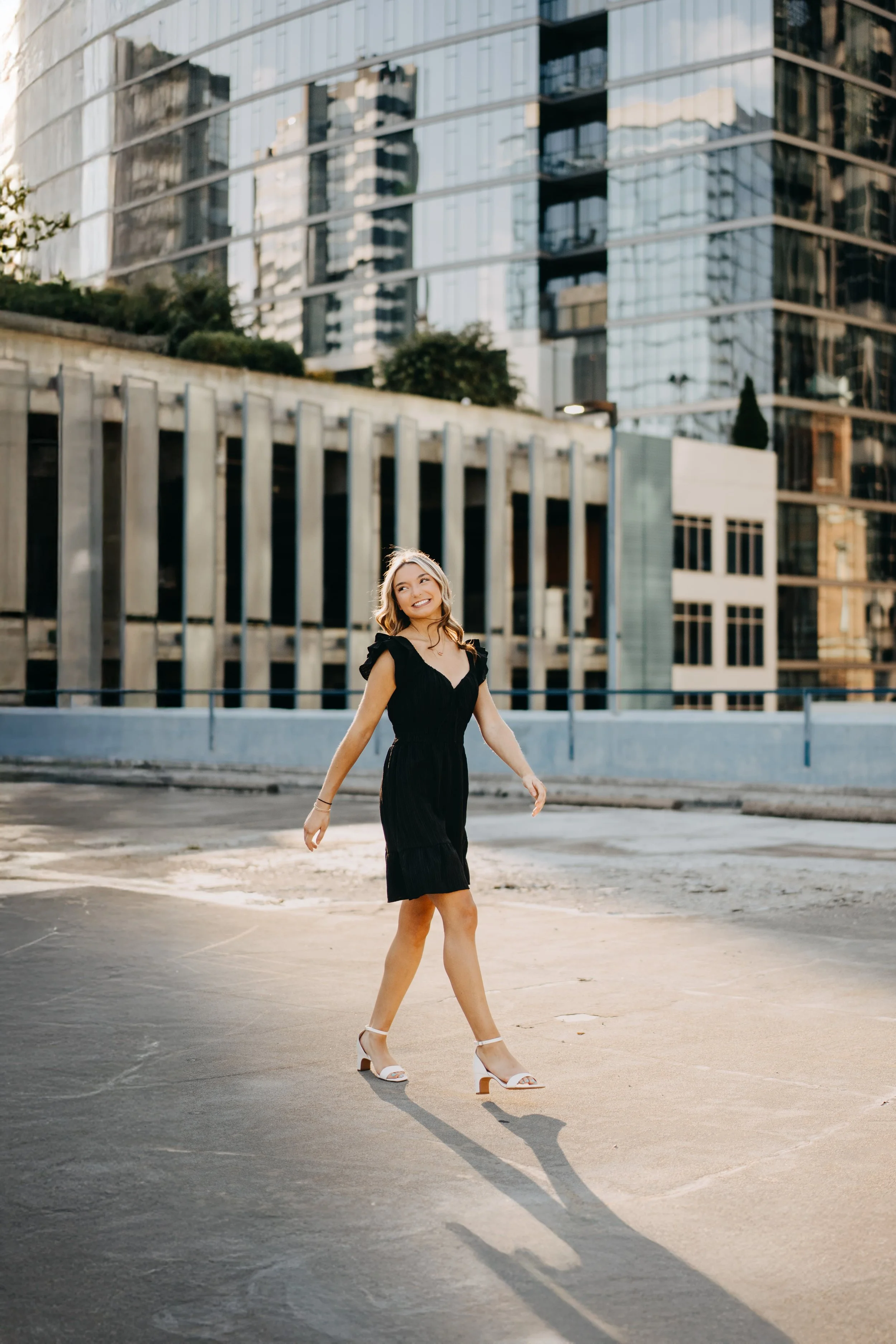 A woman in a black dress and white high heels walking outdoors on a city street, with modern glass buildings in the background.