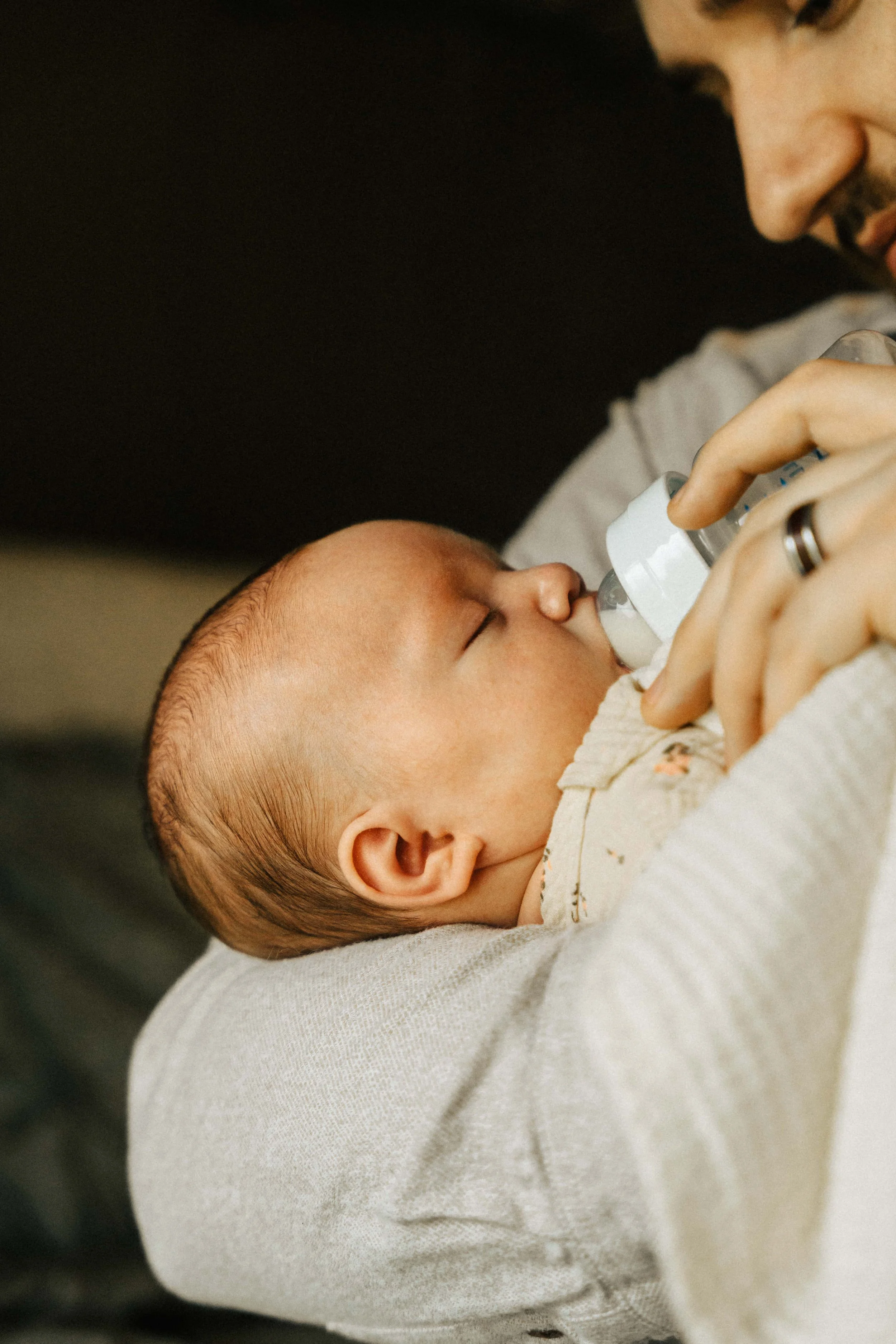 A baby is being fed with a bottle by an adult, possibly a parent, who is holding the baby and supporting its head. The baby is lying on the adult's arm, and the focus is on the baby's face and the bottle.