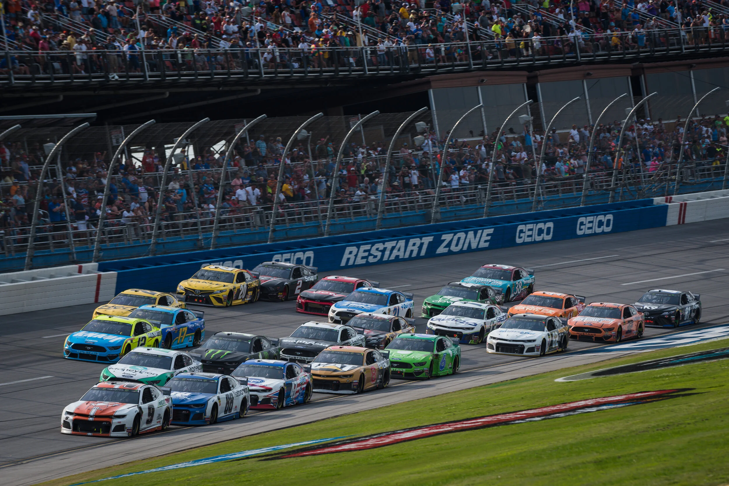 Multiple race cars on a NASCAR track during a race, with spectators watching from the stands and a blue and white wall marked with the words 'RESTART ZONE' and 'GEICO'.