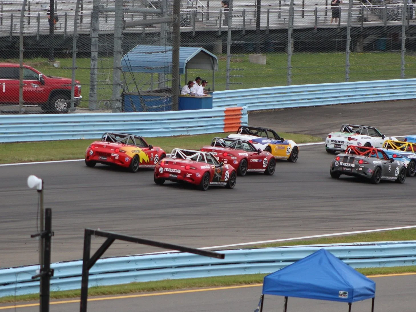 Multiple small race cars with roll cages navigating a turn on a race track, with personnel and a blue tent nearby.