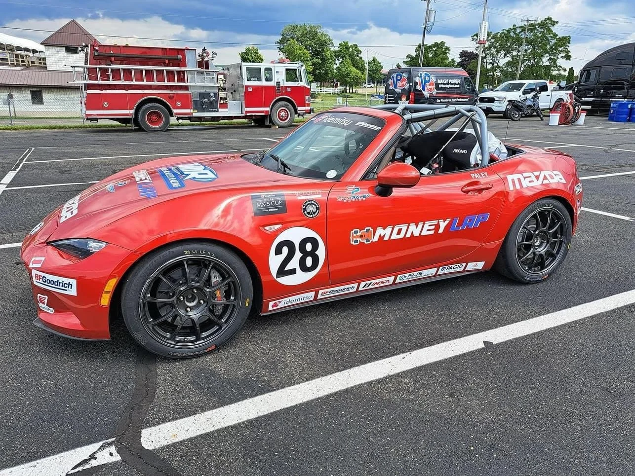 Red Mazda MX-5 race car with the number 28, sponsor logos, and a roll cage, parked in a lot with fire trucks and race team vehicles in the background.