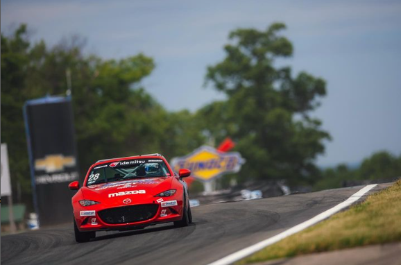 Red Mazda race car on a race track, approaching a turn, with trees and a blurred sign in the background.