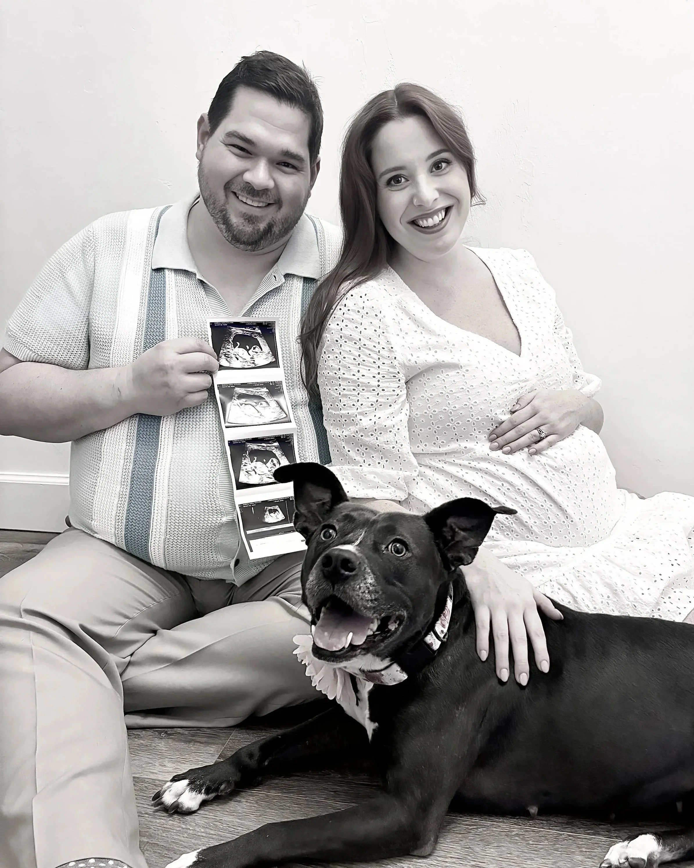 A black and white photo of a pregnant woman with a man and a dog, smiling and holding up a strip of ultrasound photos
