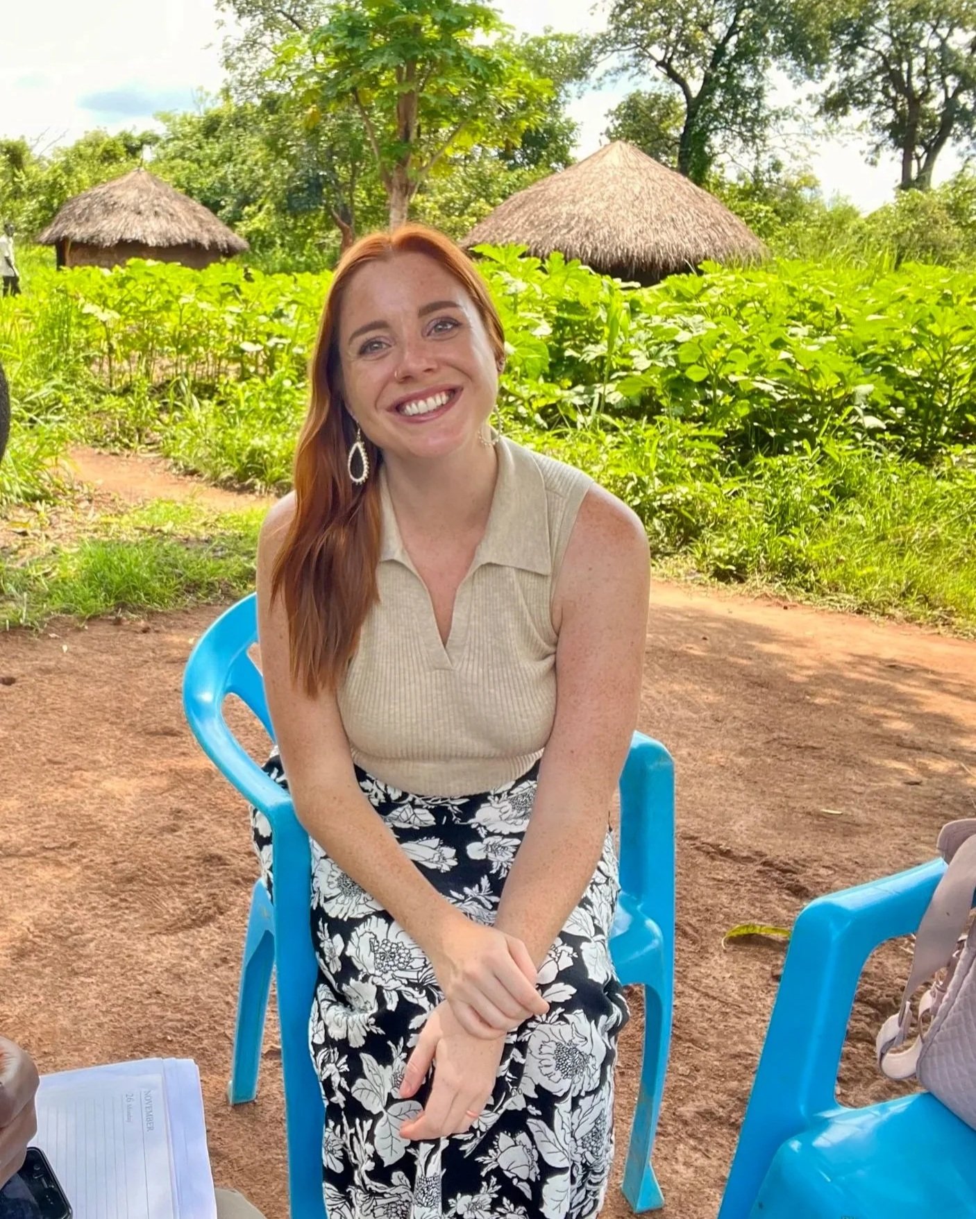 A picture of a white woman with red hair sitting in a blue chair and smiling in front of a scene of trees and huts