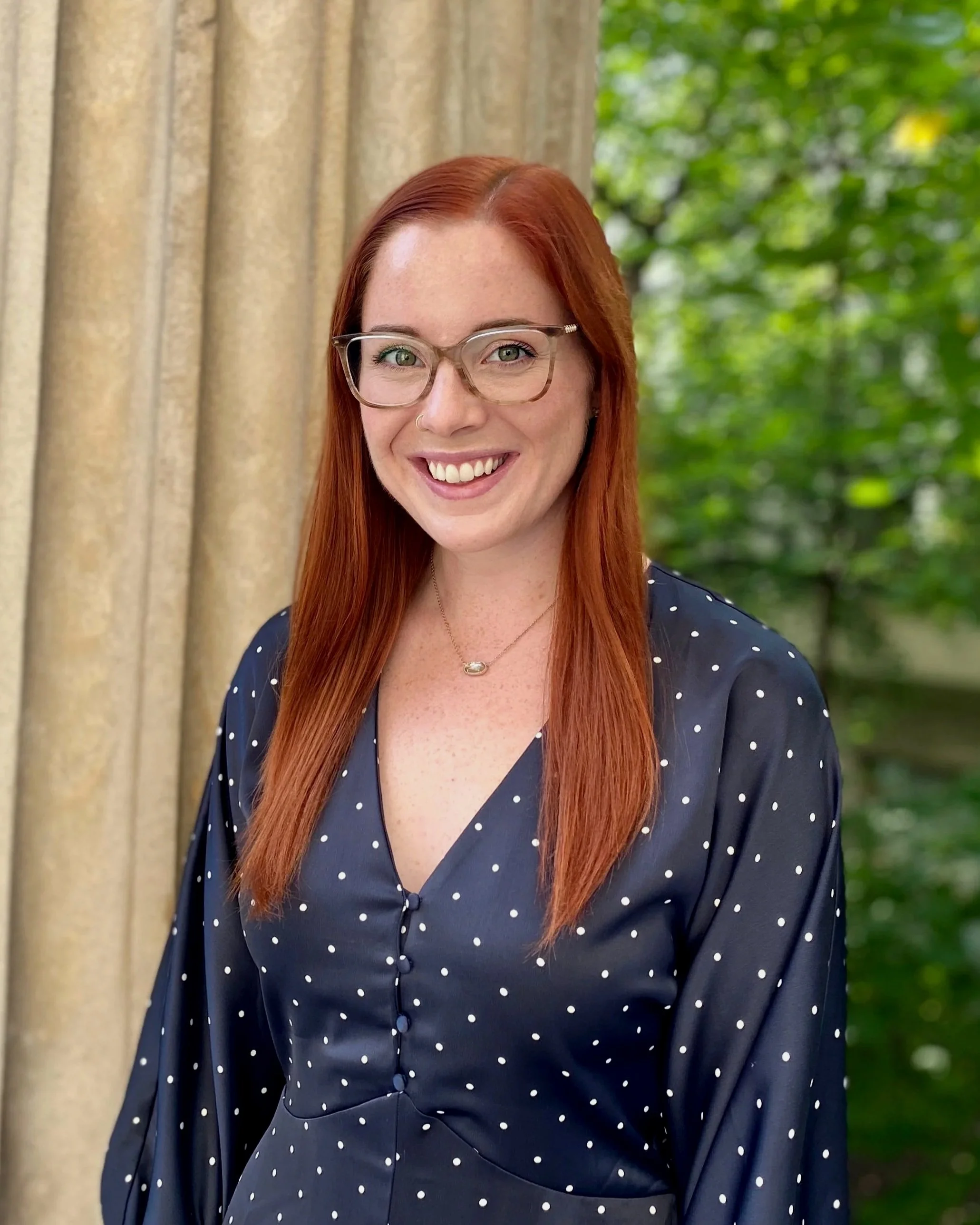 A headshot of a white woman with red hair and glasses smiling in front of a grey column
