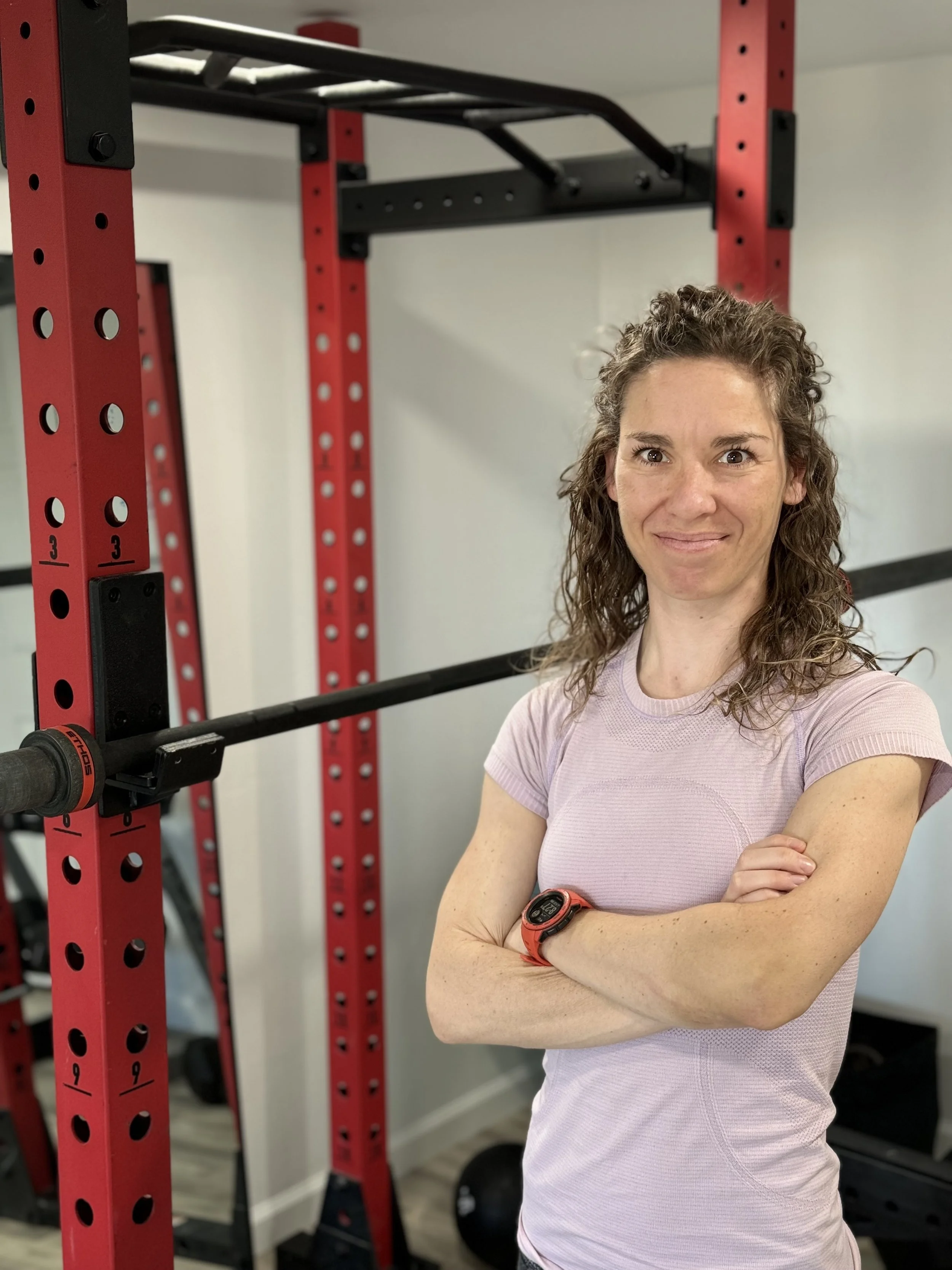 A woman with curly brown hair standing in a gym next to a red and black power rack, crossing her arms and smiling at the camera, wearing a light pink athletic shirt and a red watch.