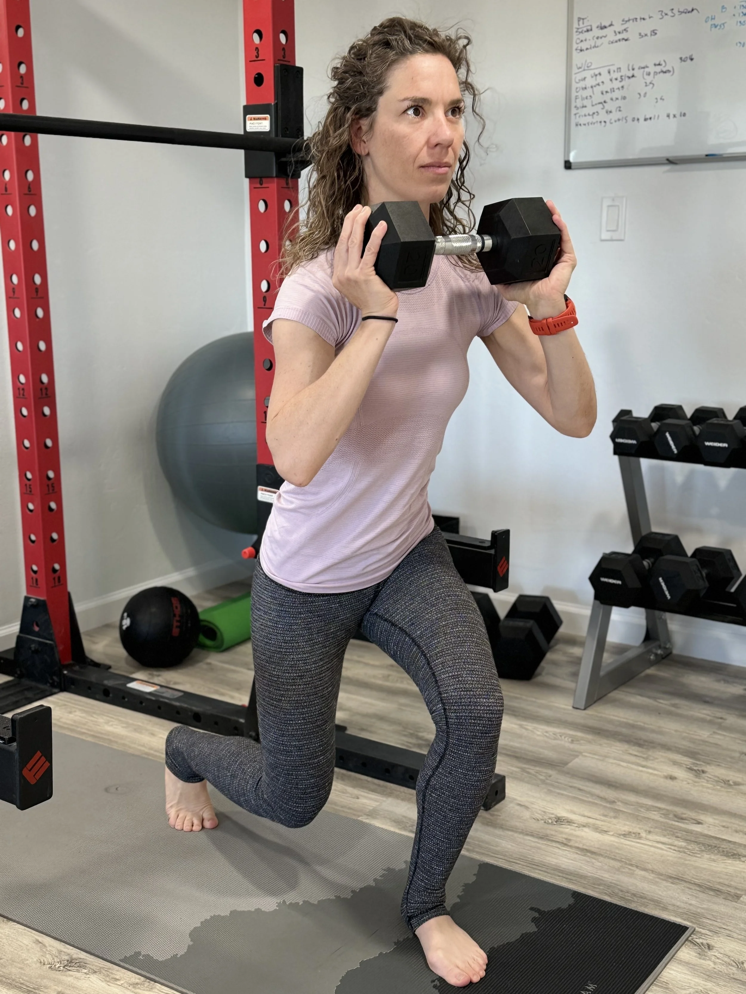 Woman in workout clothes practicing a lunge exercise while holding dumbbells at home gym.