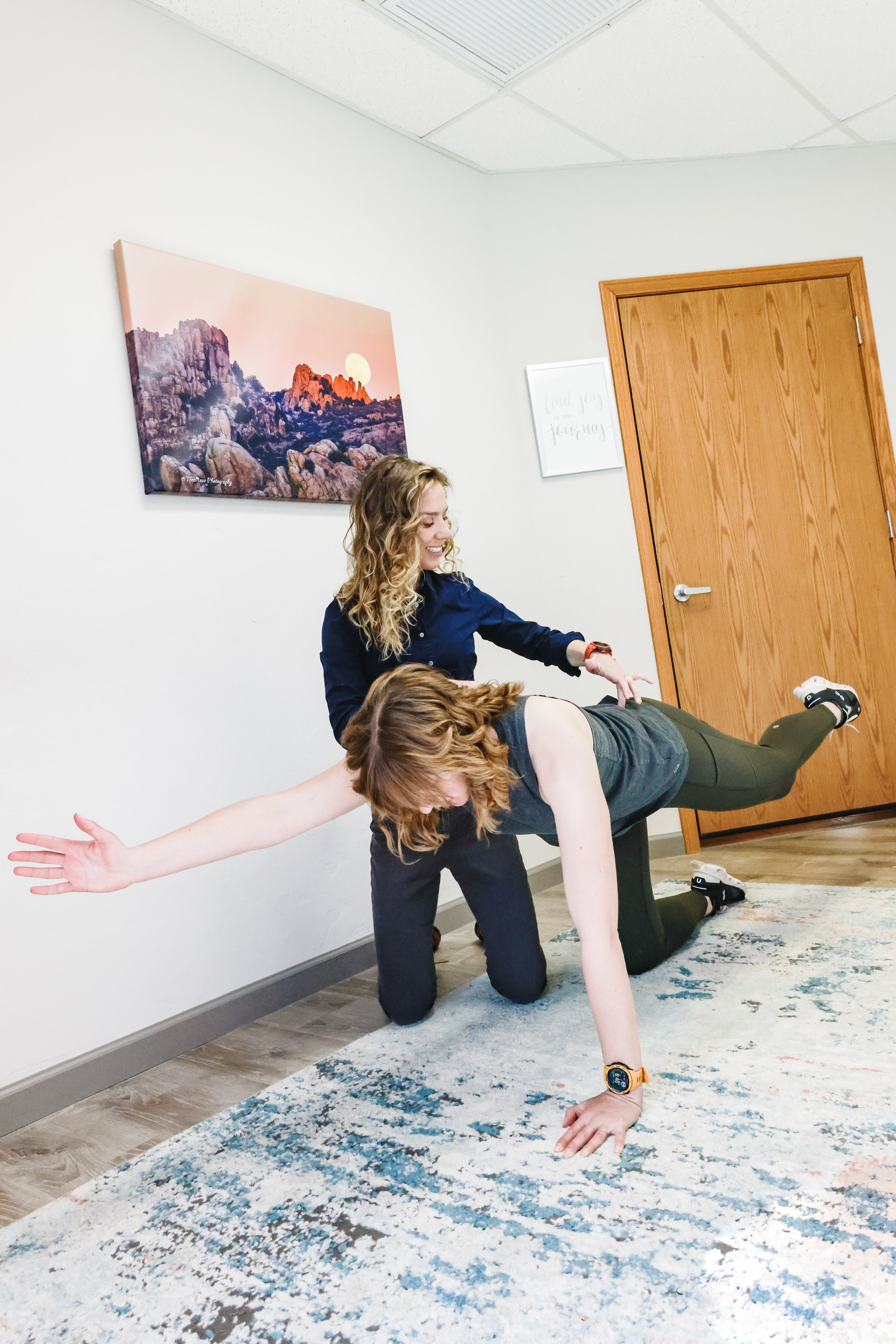 Two women exercising, one is supporting the other in a plank position with a hand on the floor and her leg raised, in a room with a wooden door, framed wall art, and a landscape painting on the wall.
