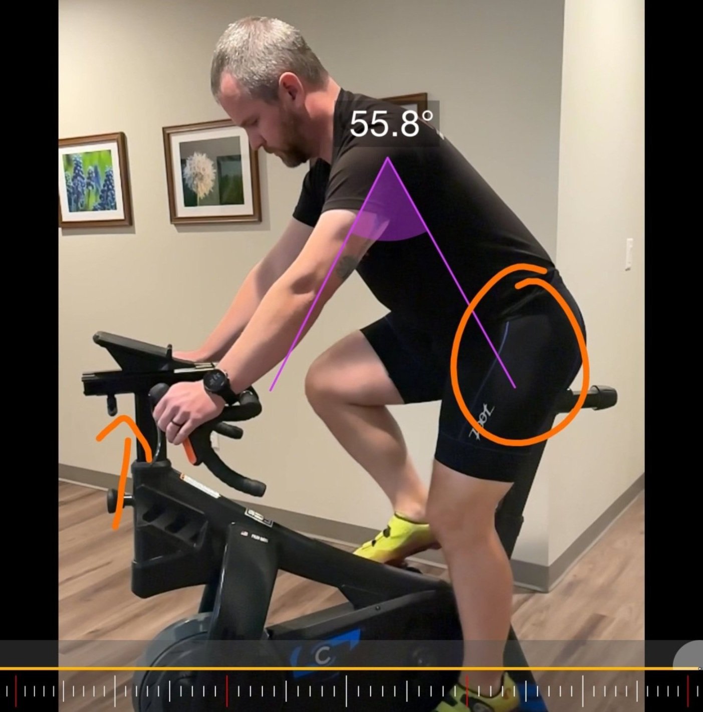 A man riding a stationary indoor bike, wearing a black shirt and shorts, with measurements and angles overlaid indicating a 55.8° back angle, and orange circles highlighting leg and hip positions.