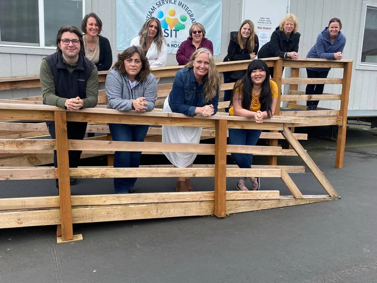 Group of ten women and one young woman are gathered around a wooden ramp outside a building. Some are standing behind the ramp and some are leaning on it. They are smiling and looking at the camera. There is a sign on the building with the logo and text for SIT, emphasizing team service, integration, and embracing the community.