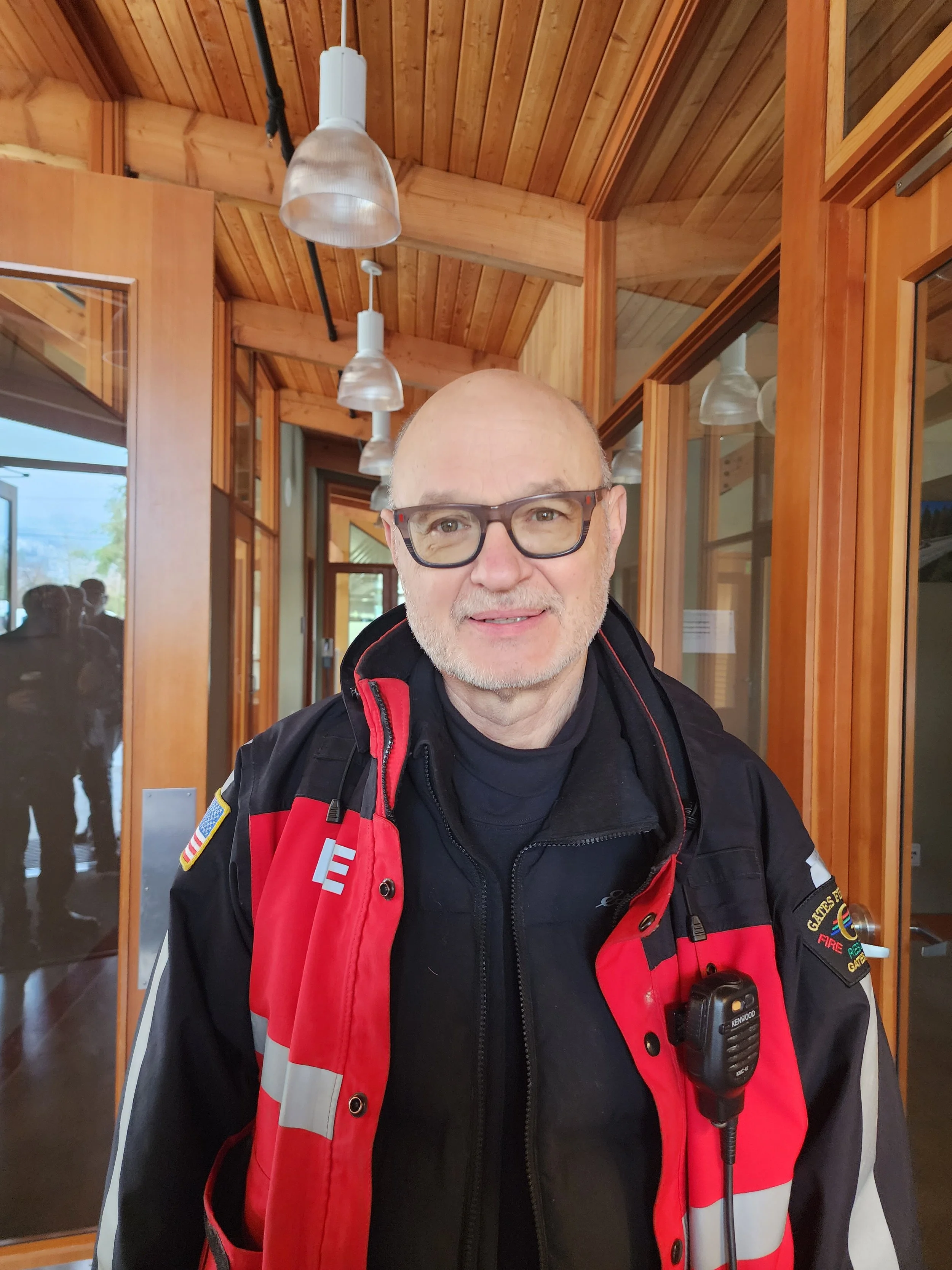 A man with glasses wearing a firefighter uniform and a red vest, standing indoors with wooden walls and glass doors.