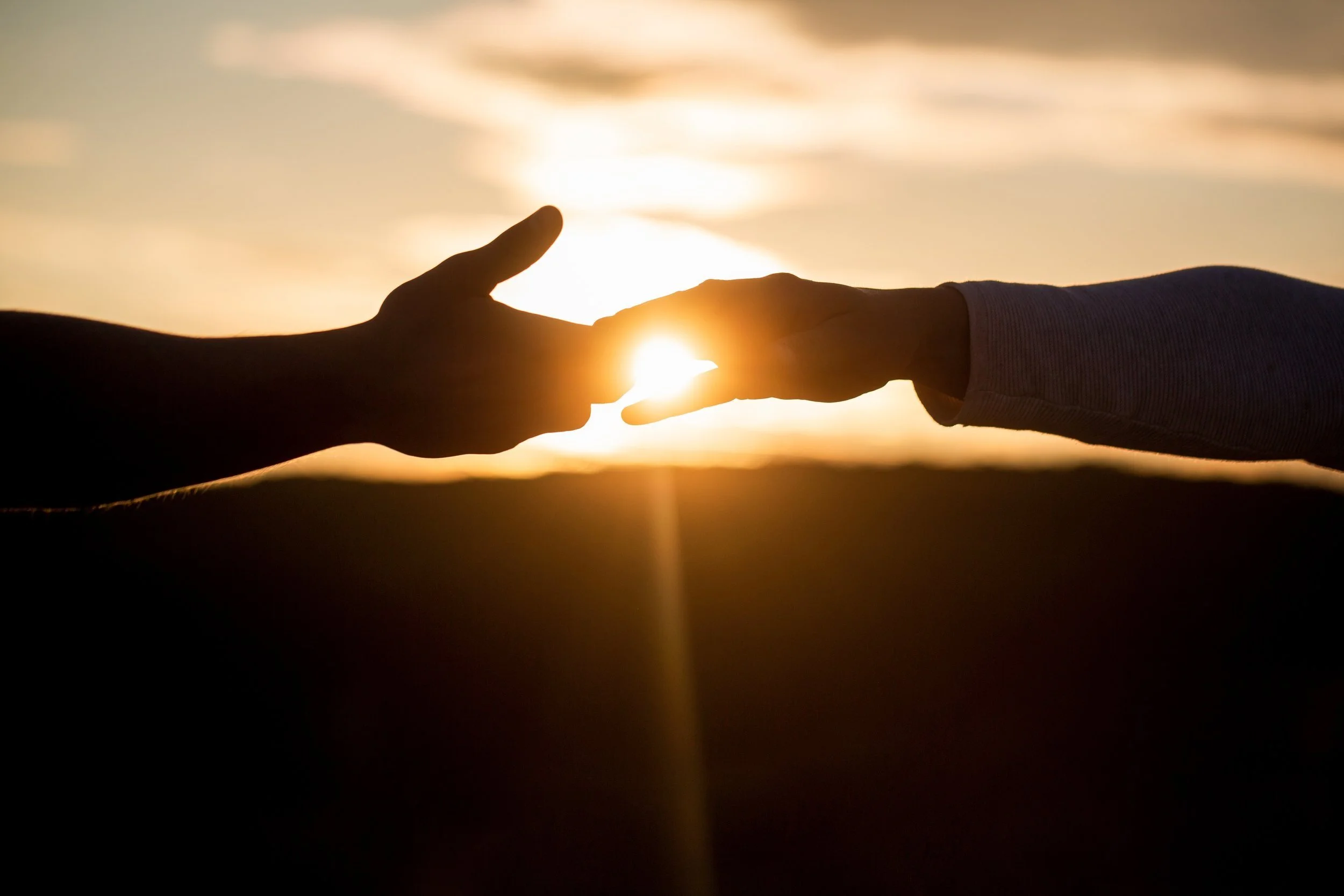 Two hands reaching towards each other with a sunset or sunrise in the background.