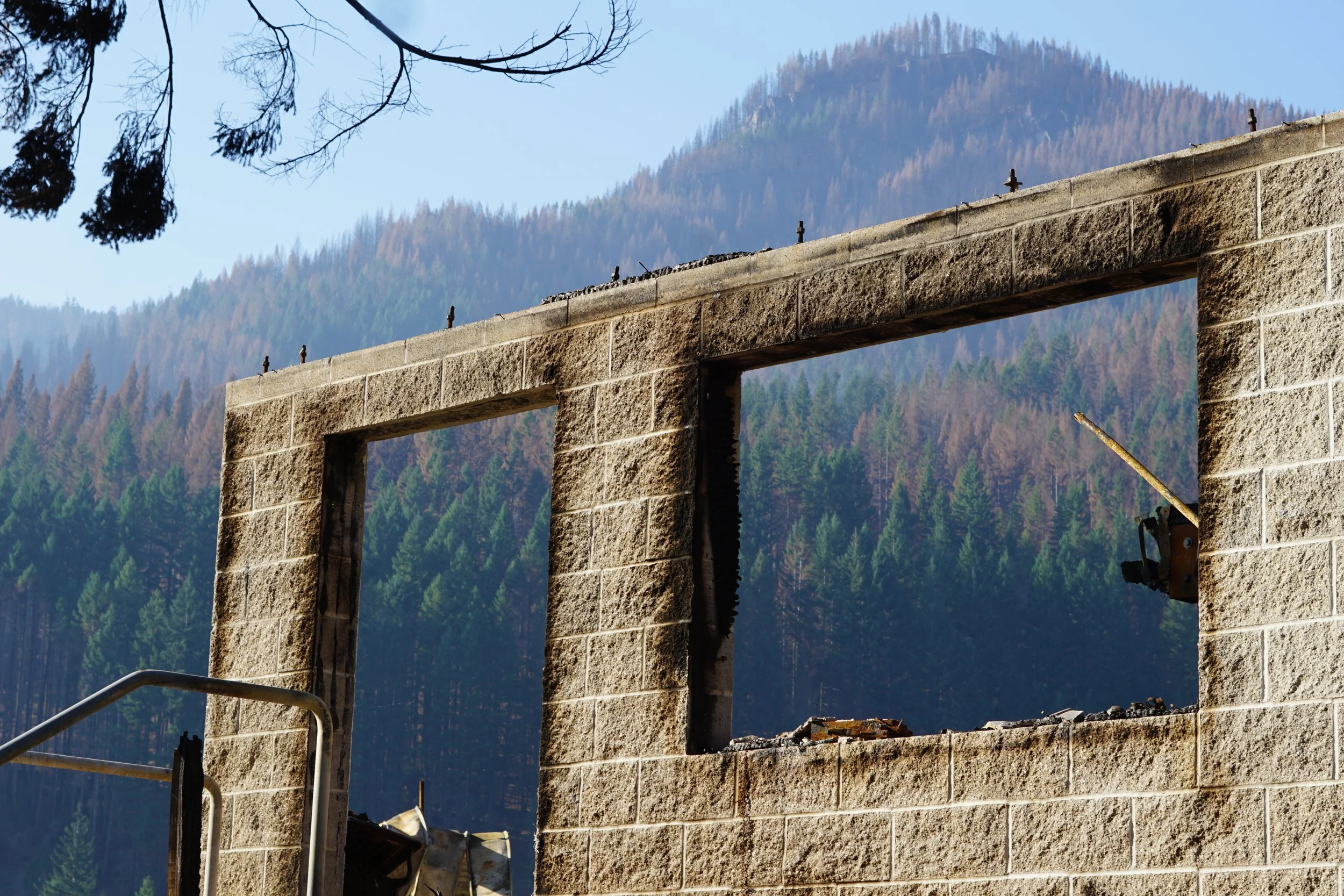 Partial view of a stone building with two large rectangular openings, set against a backdrop of forested mountains. There are some construction materials and a metal railing visible.