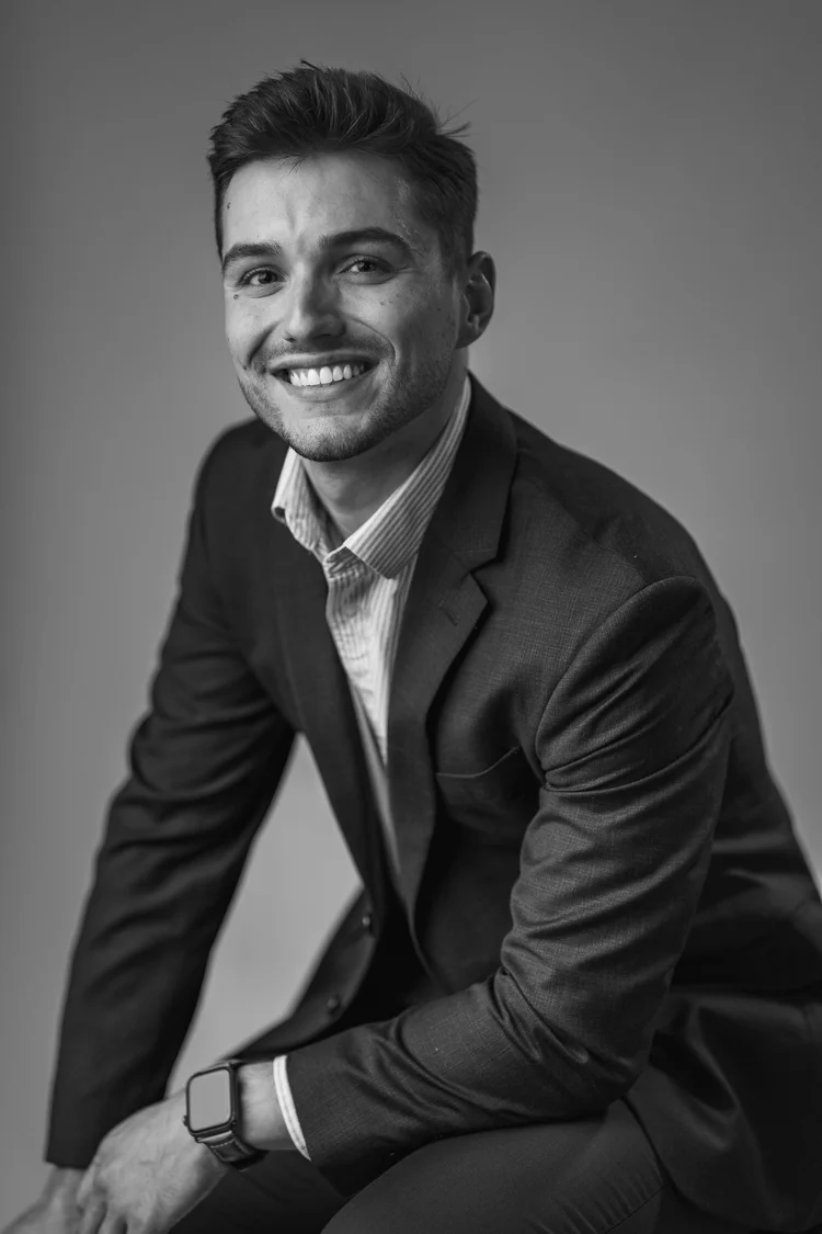 Black-and-white headshot of a smiling young man in a dark suit jacket and striped shirt, leaning forward on a stool, Lake Charles