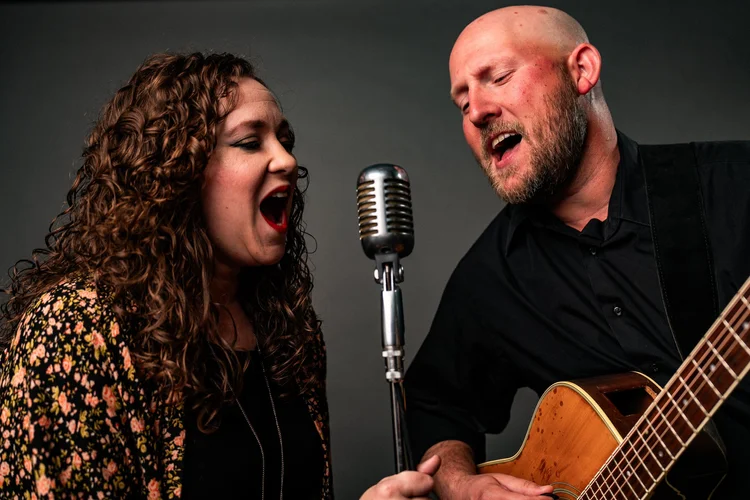 Musical duo singing together into a vintage microphone, a curly-haired woman in a floral top and a bearded man playing acoustic guitar, band promo photo, Lake Charles