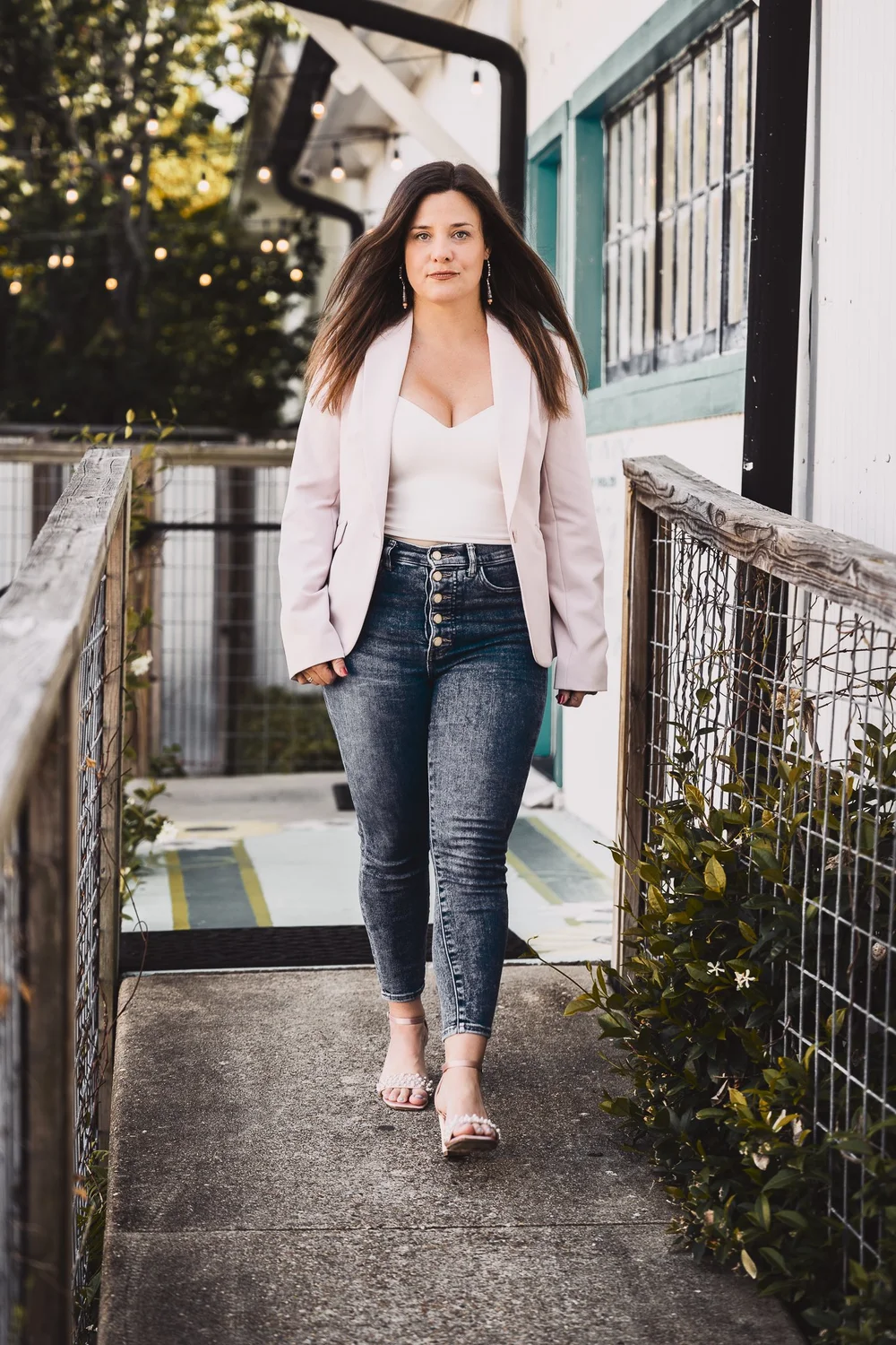 Katy Rozas in a light pink blazer and jeans walking down a plant-lined walkway in Lake Charles, branding portrait by Wildwoods Creative