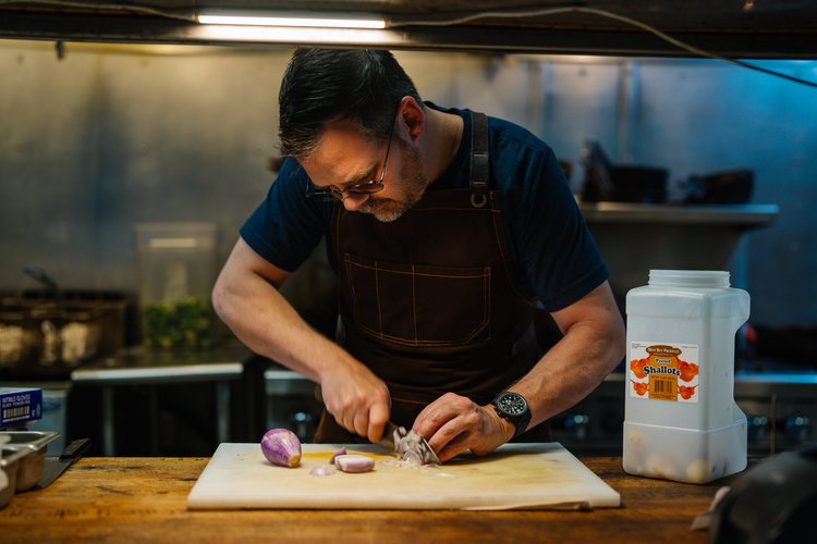 Chef in glasses and a brown apron slicing shallots on a cutting board in a restaurant kitchen, restaurant branding photo, Lake Charles