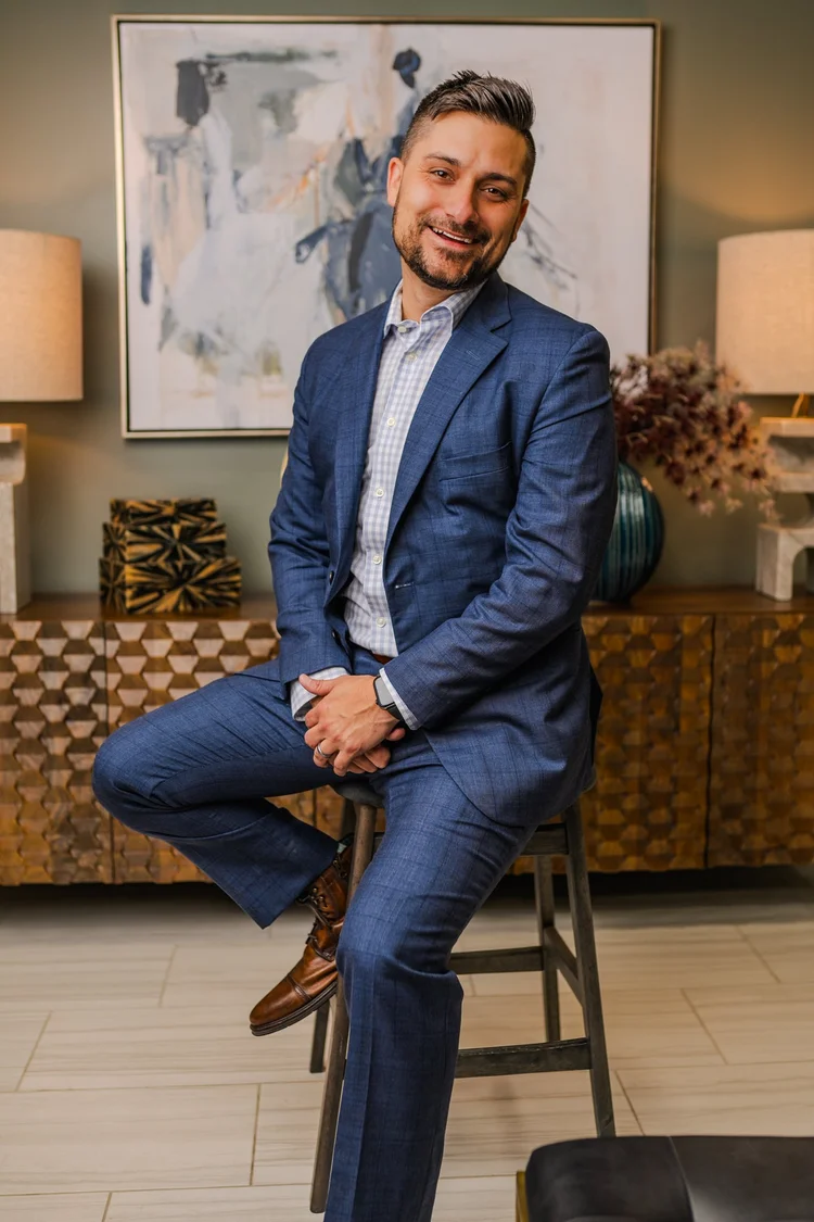 Smiling man in a blue plaid suit sitting on a wooden stool in a styled lounge with abstract art behind him, branding portrait, Lake Charles
