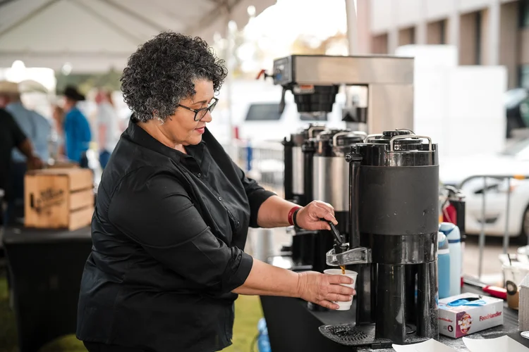 Woman with curly gray hair in a black chef's shirt pouring coffee from a steel urn into a paper cup at an outdoor event coffee station, Lake Charles