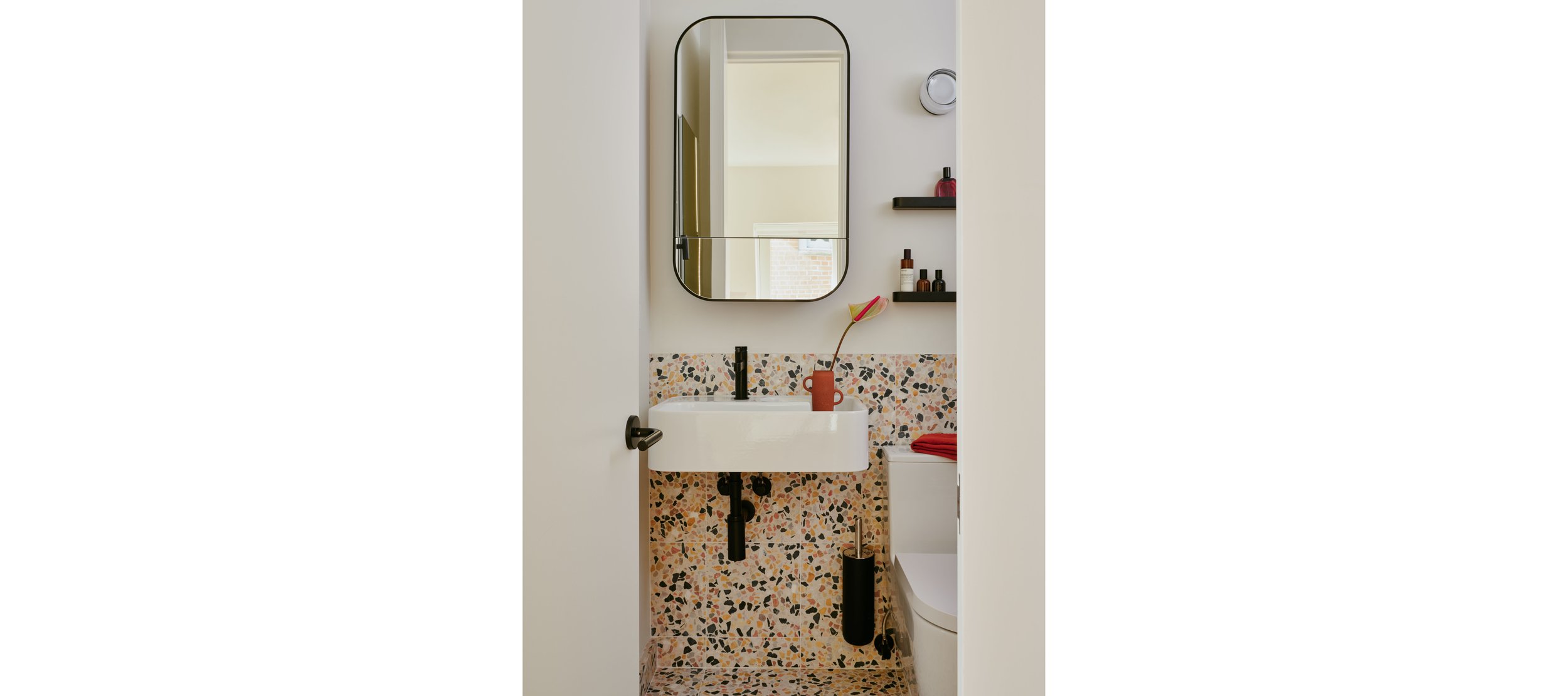Small bathroom with a white sink, terrazzo wall tiles, a wall mirror, and black fixtures. Shelves with decorative bottles and a flower in a small vase on the sink.