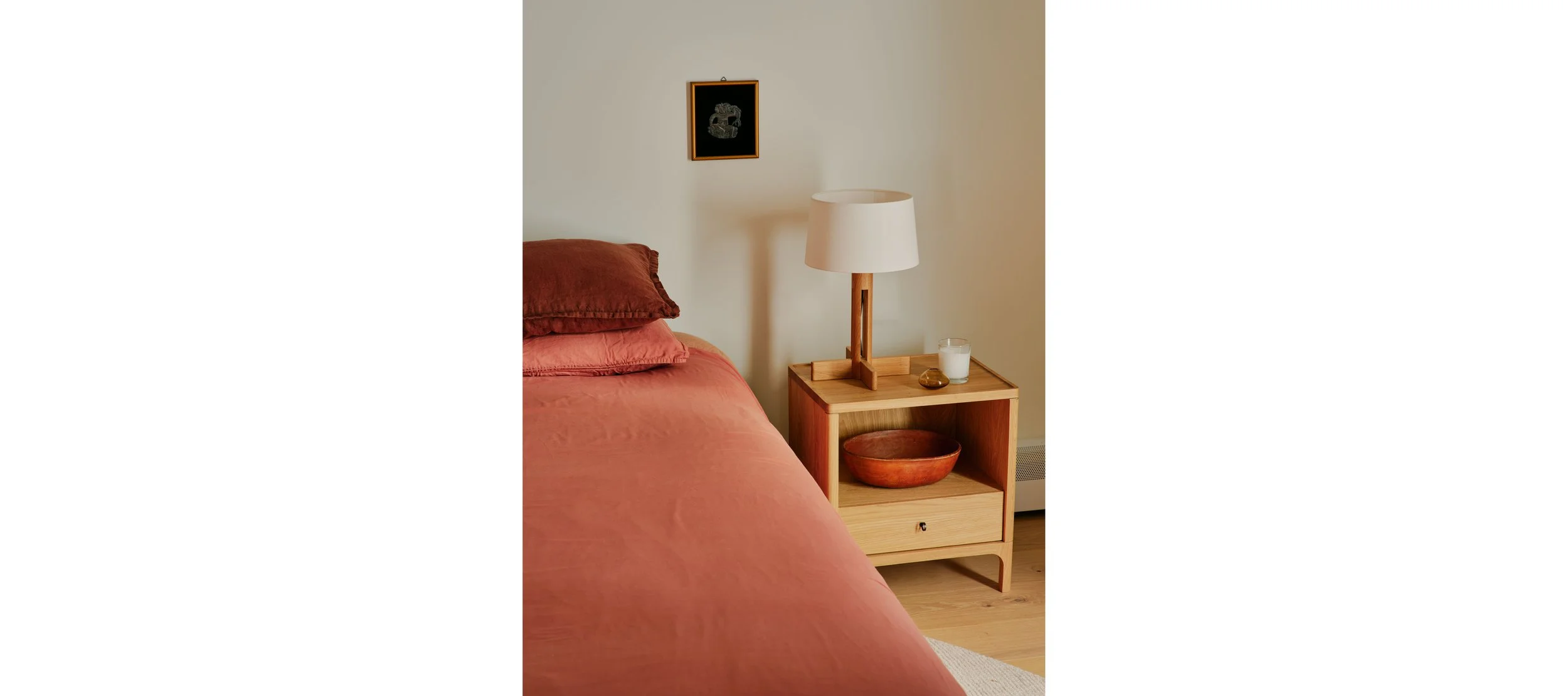 Close-up of a bedroom corner with a bed featuring peach-colored sheets and rust-colored pillows, a wooden nightstand with a drawer, a table lamp with a white shade, a glass of milk, a small decorative object, a wooden bowl on the lower shelf, and a s
