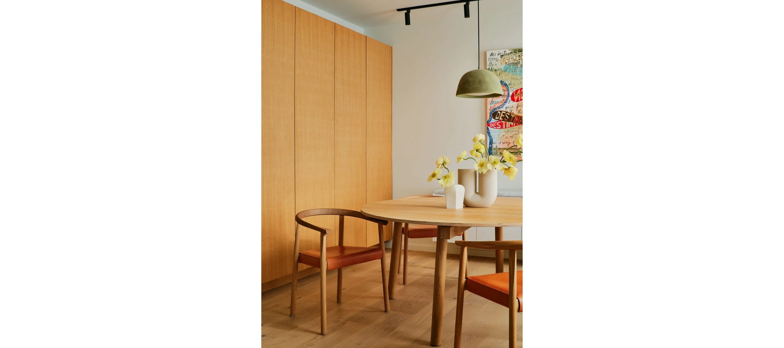 A dining room with a wooden round table, two chairs with orange cushions, a white vase with yellow flowers, a green pendant light, wooden cabinetry, and colorful abstract artwork on the wall.