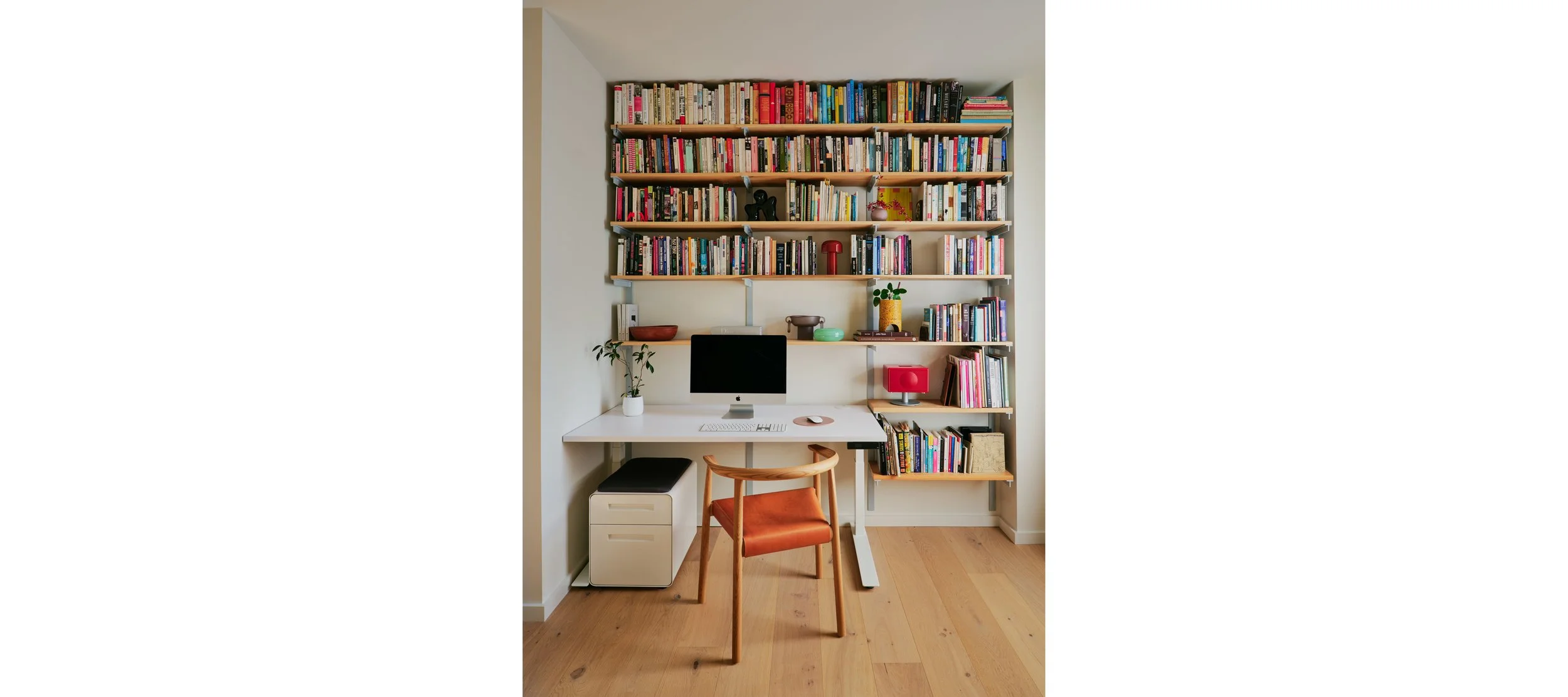 A home office with a white desk, an iMac computer, a wooden chair with an orange cushion, and a large bookshelf filled with books and decorative items.