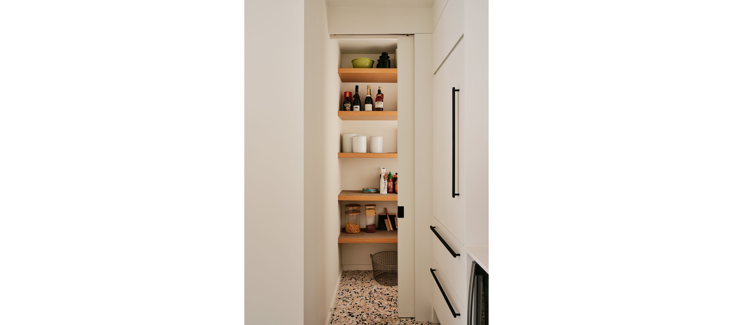 Narrow kitchen pantry with wooden shelves holding bottles, bowls, candles, jars, and seasonings, on a terrazzo floor.