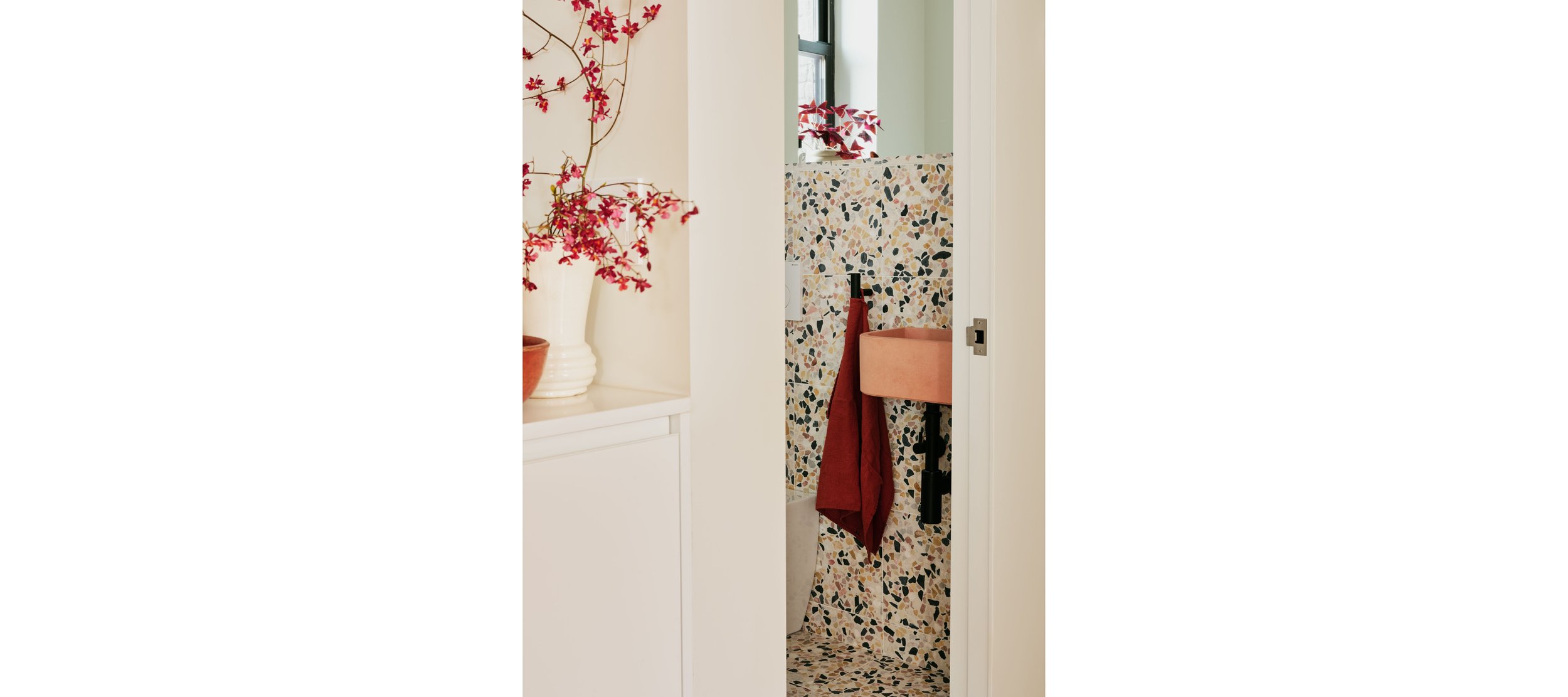 Partial view of a bathroom with terrazzo tile walls and floor, a small peach sink, and a red towel hanging, with a window and decorative plants in the background.