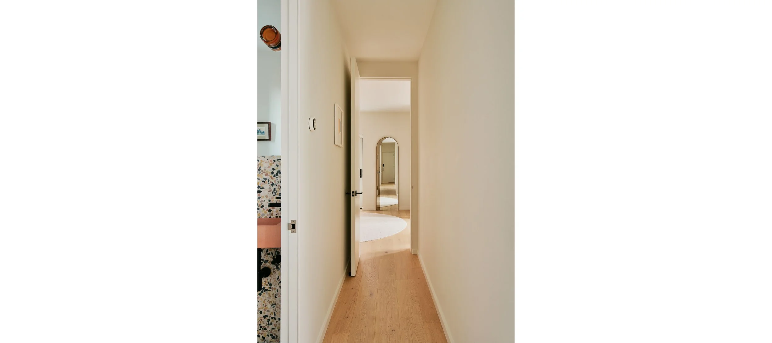 View of a narrow hallway with wooden floors leading to a room with a round white rug and a mirror on the wall.