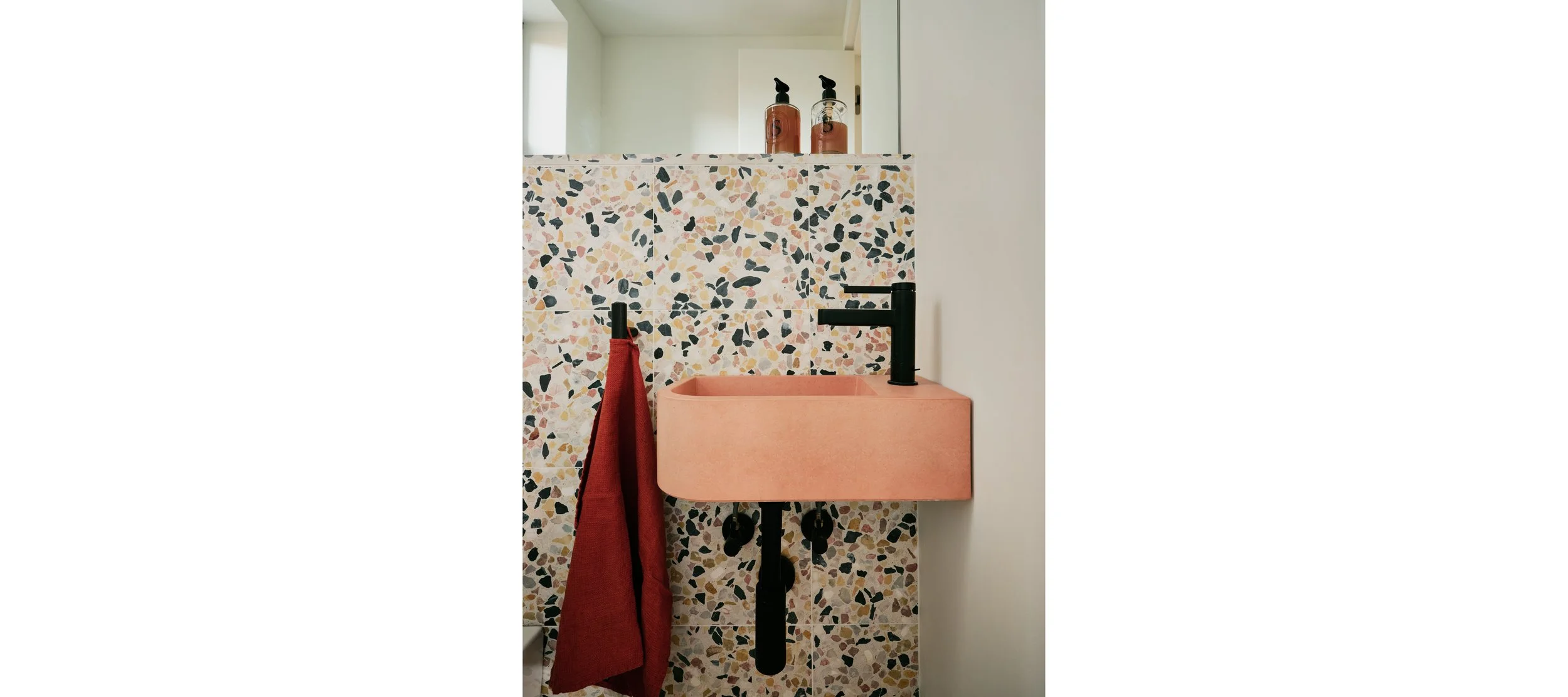 Pink rectangular bathroom sink with black faucet, a red towel hanging on a black hook, terrazzo wall behind the sink, and two soap dispensers on a shelf above.