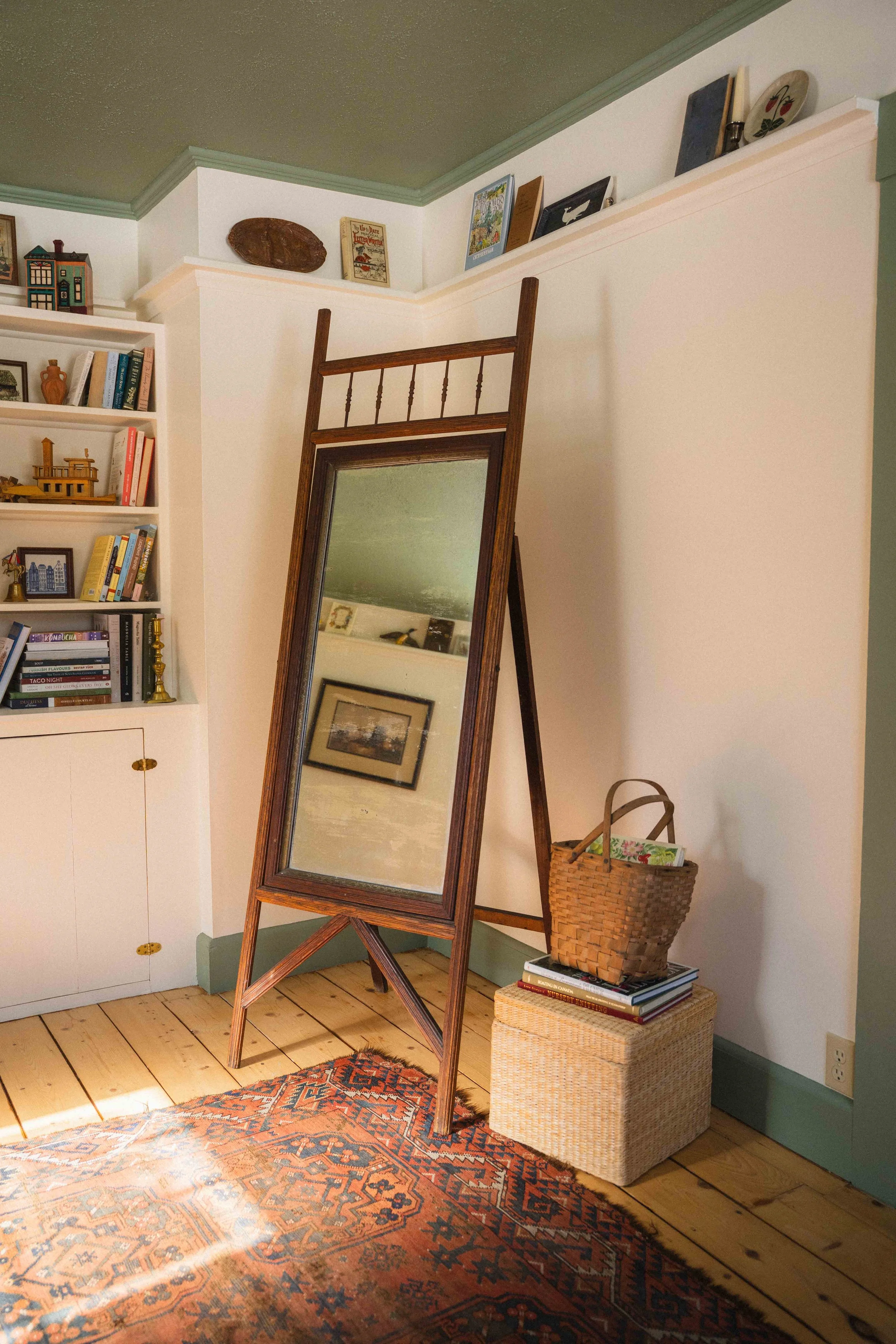 an antique wood framed cheval mirror in a cottagecore library with a woven basket, wicker footstool, a turkish rug, and colourful bookshelf with a styled plate rail, cream walls with green ceiling and trim. historic home design
