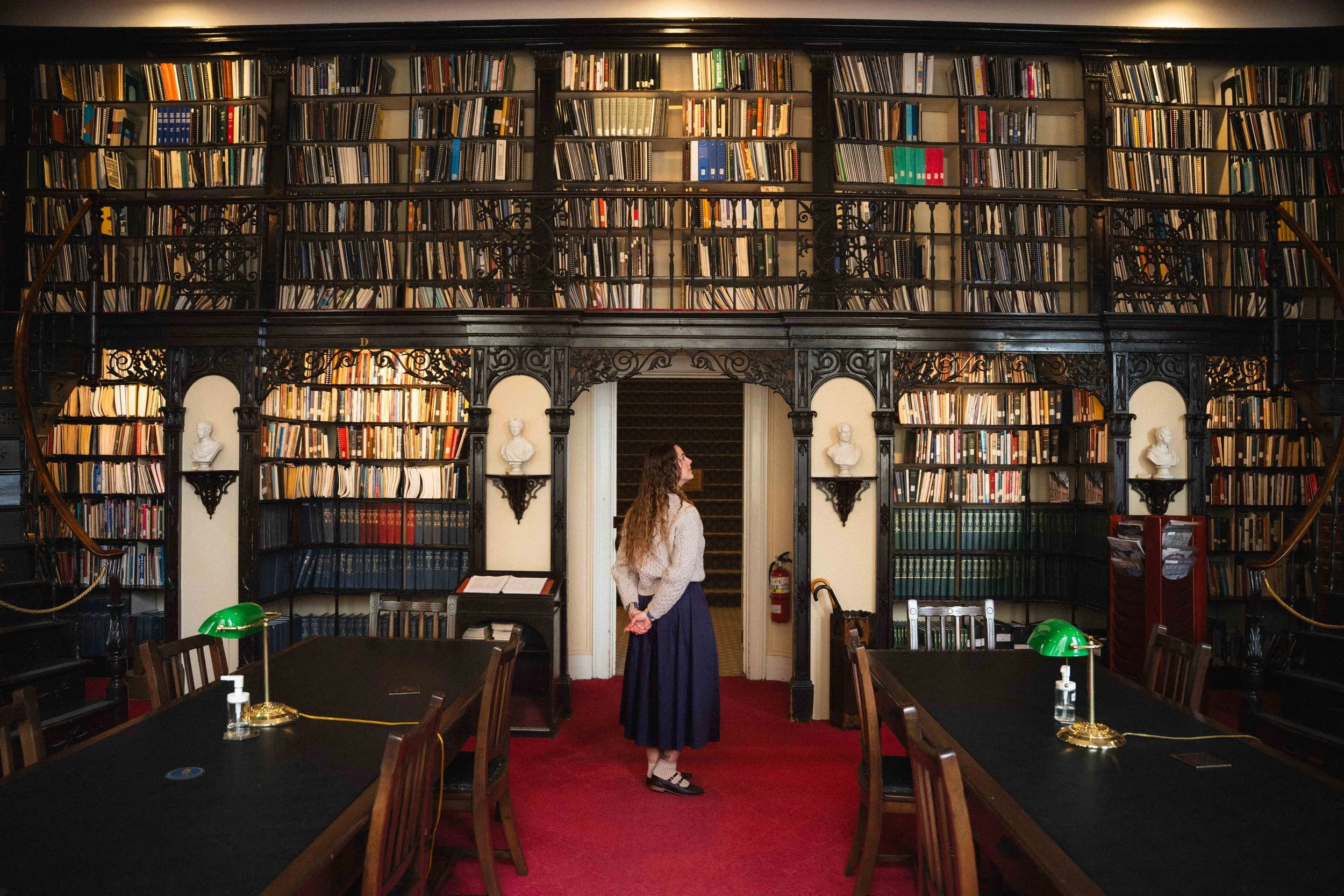 a girl standing in the centre of a historic library looking up at a two story wall of books, wearing a dark academia inspired outfit with midi skirt, irish wool sweater, and socks with mary janes. The library has busts and old books with vintage desk