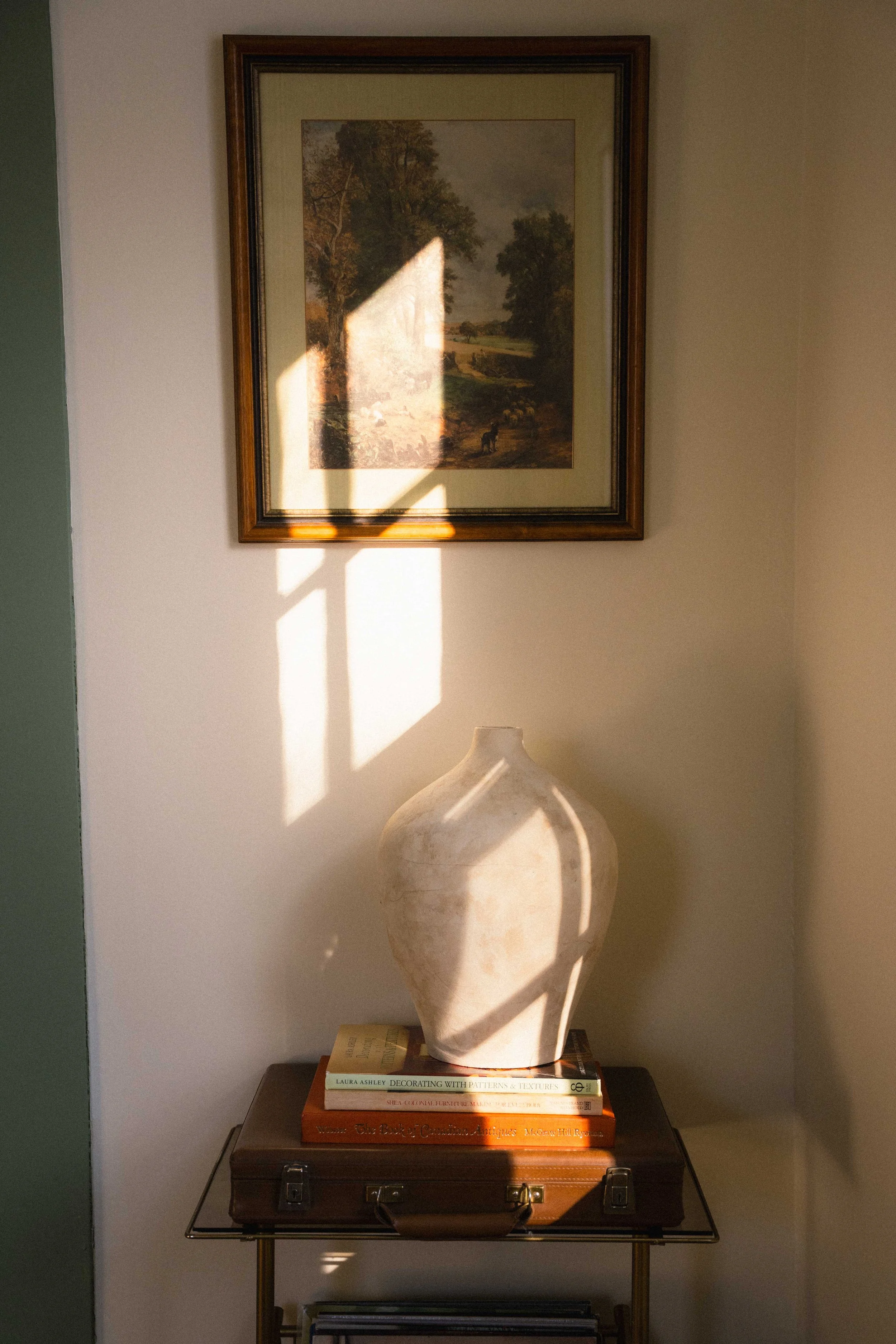 sunlight cascading over a wall with a cottagecore framed painting on a cream wall with green trim, a ceramic vase on a stack of colourful home design books - romantic interiors