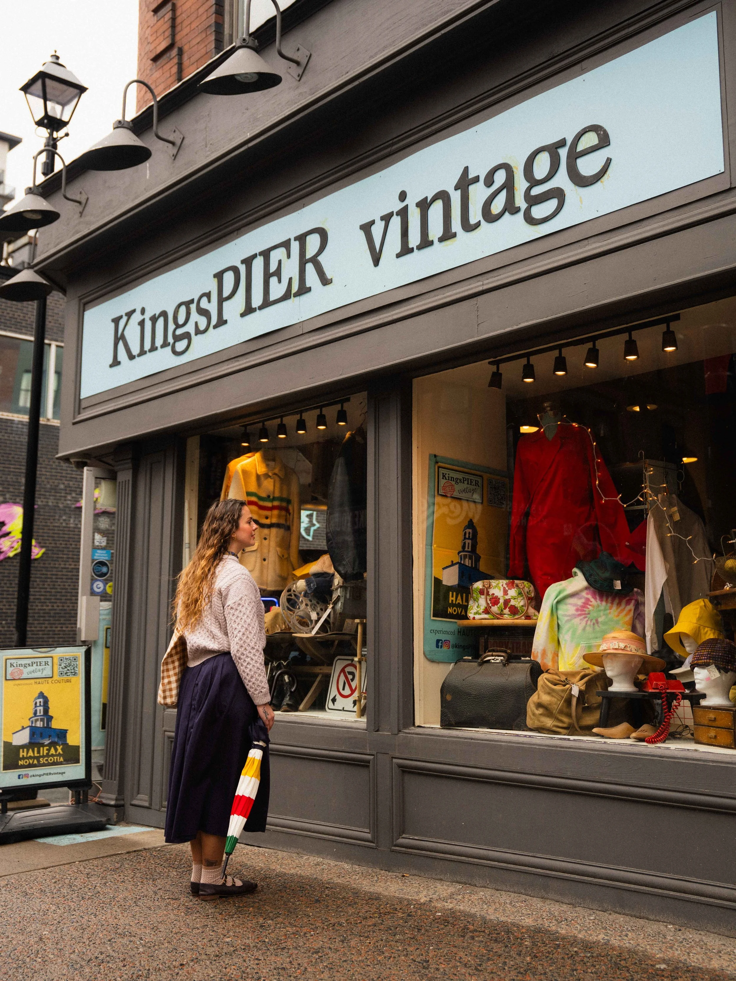 a girl looking at the window display of a vintage clothing store called KingsPIER vintage, on a rainy day, holding a Hudson's Bay striped umbrella and wearing an irish knit sweater and skirt in downtown Halifax, Nova Scotia shopping for clothes