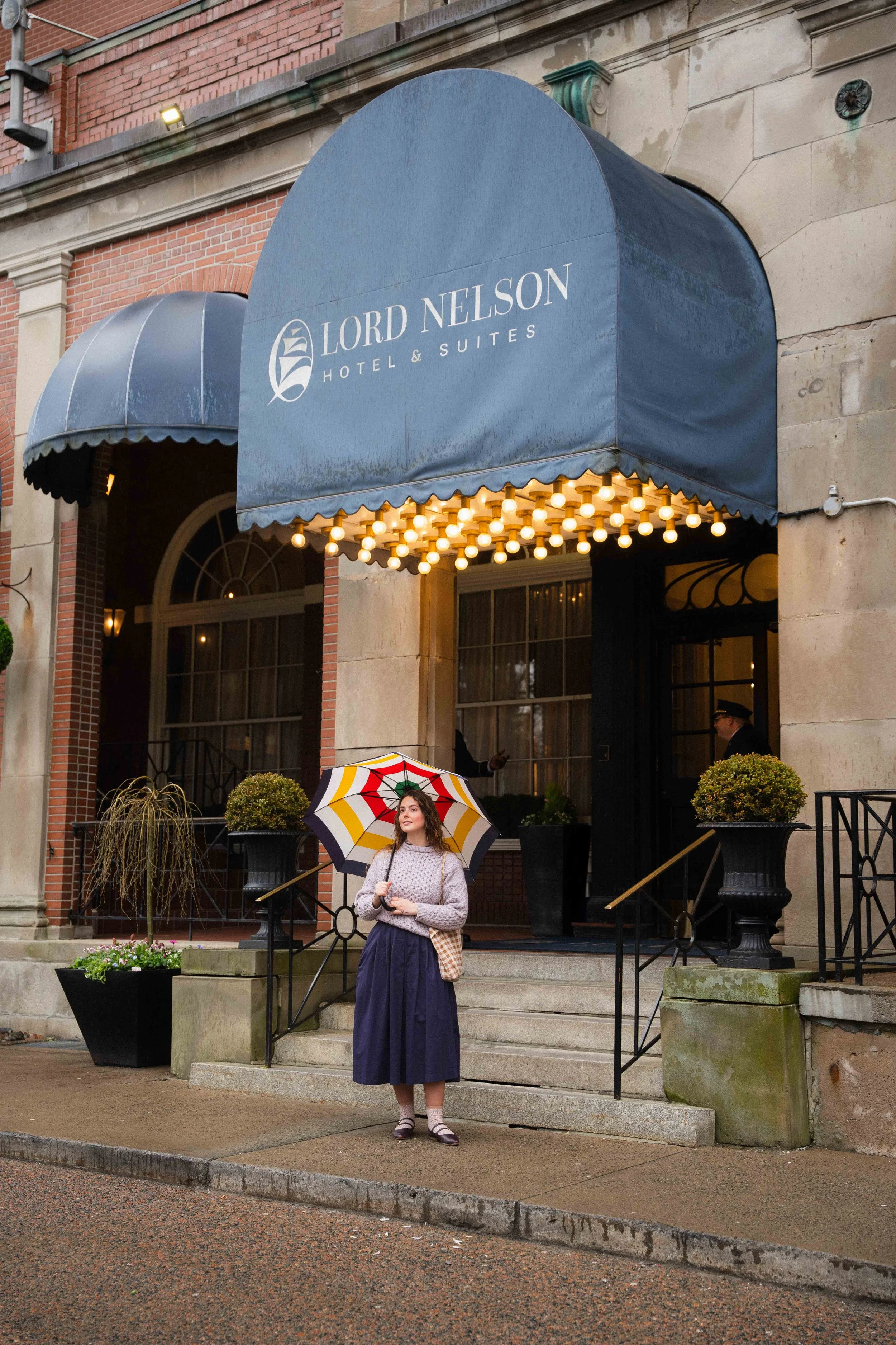 outside the historic railroad hotel, Lord Nelson Hotel & Suites. A girl wearing a dark academia outfit with a Hudson's Bay striped umbrella wearing an irish wool sweater, pleated midi skirt, and socks with mary janes