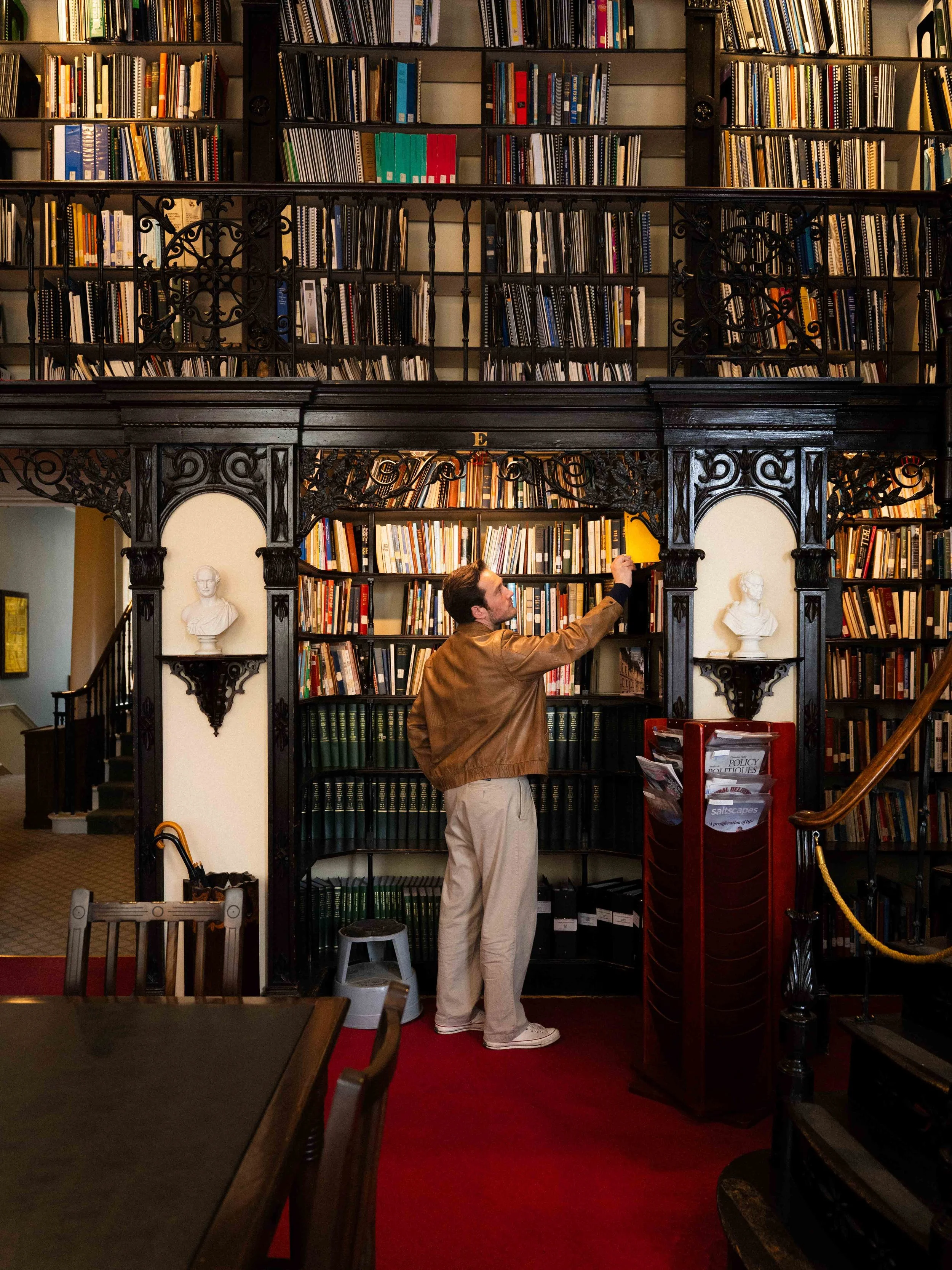 a man dressed in academia style grabbing a book off a shelf in a historic library nook