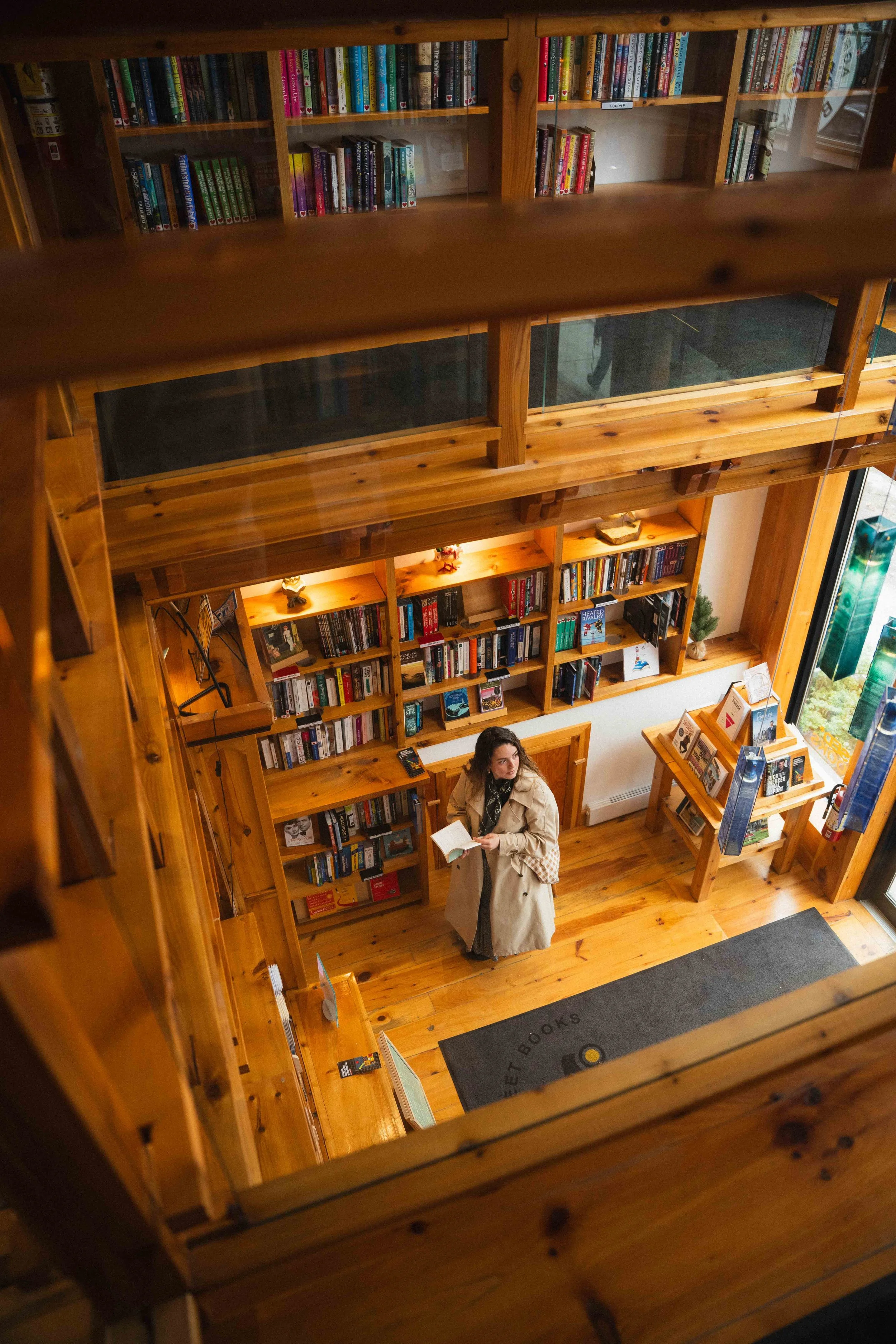 a view from a second story loft of a girl looking over her shoulder in a book store featuring warm cedar wooden shelves, the girl is wearing a long beige trench coat and neckscarf, and is holding a secondhand book