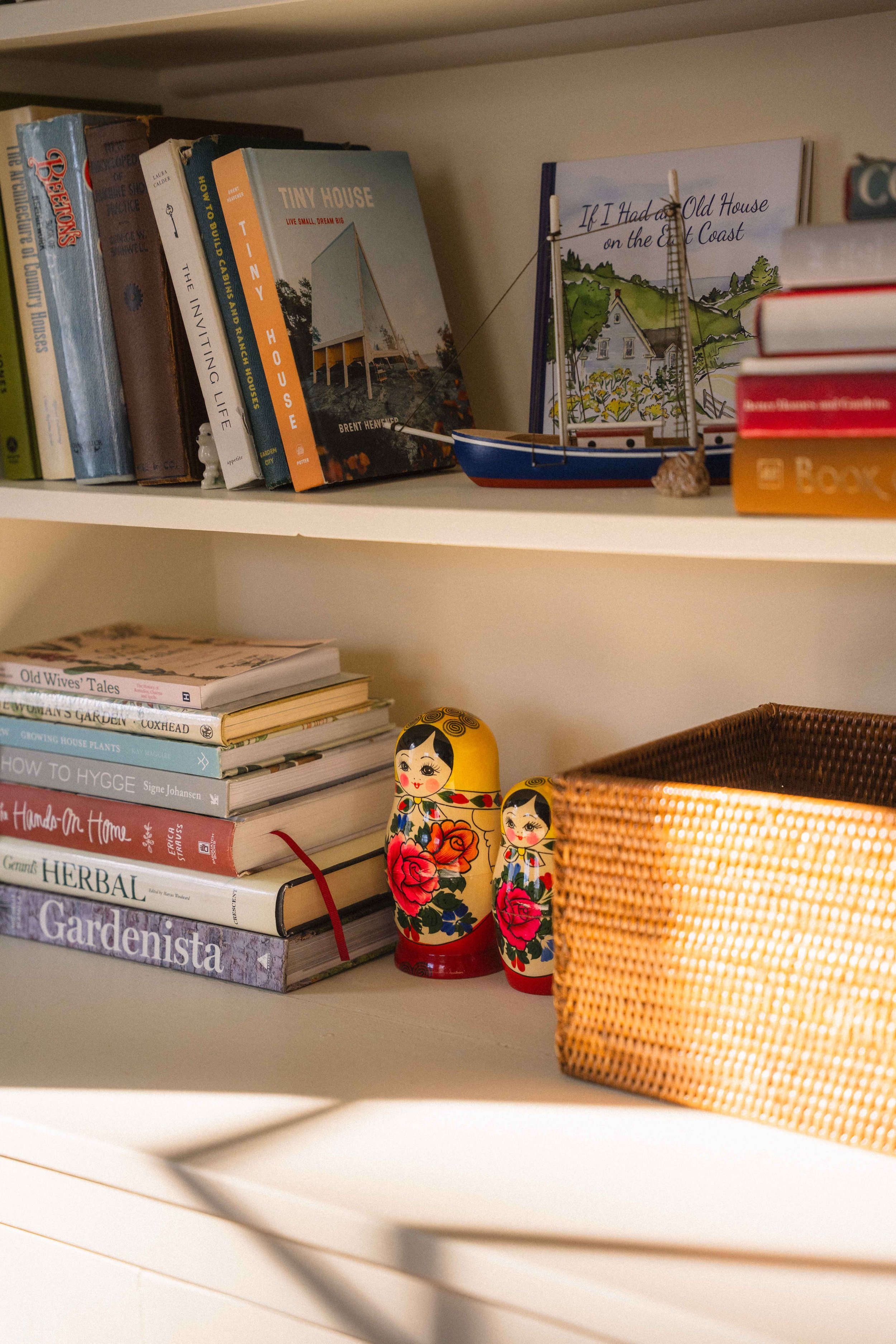 collected cream built-in bookshelf with a rattan basket, matryoshka dolls, a model boat, and book stacks with titles like Hands on Home, Gardenista, How to Hygge, If I Had an Old House on the East Coast, and Tiny House.