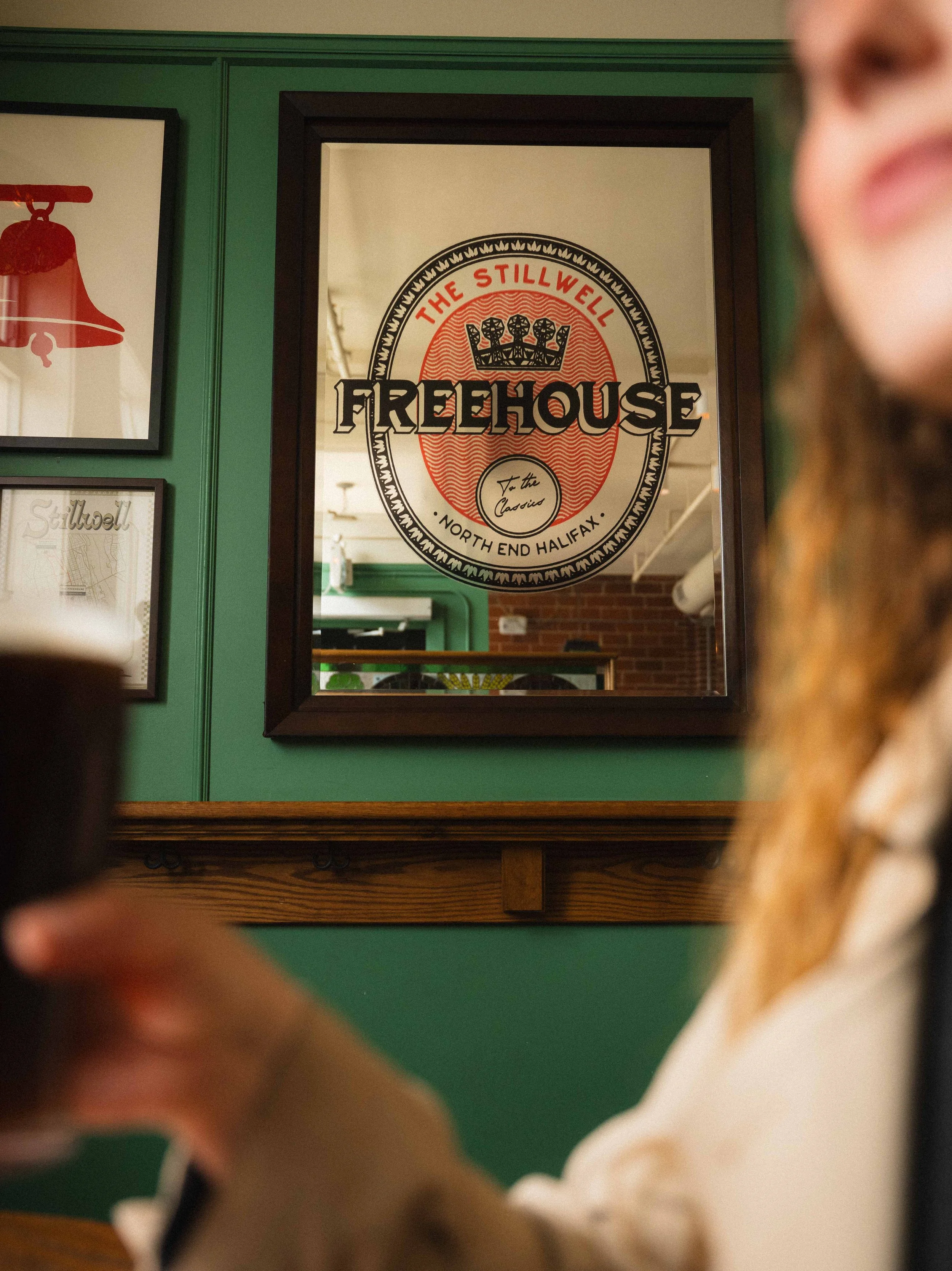 an old pub mirror sign that reads The Stillwell Freehouse to the Classics North End Halifax. A woman holding a pint of ale is blurred out in the foreground.